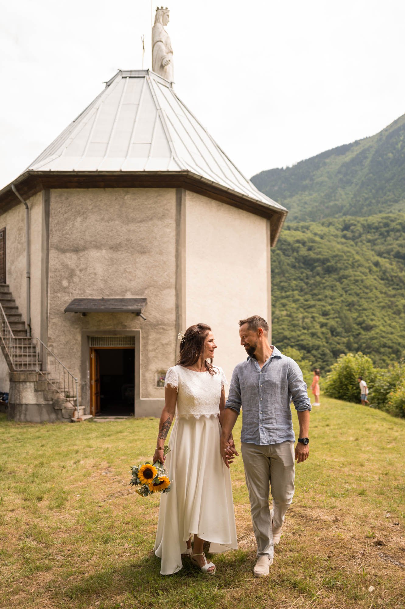 Un couple marchant main dans la main devant une petite église dans un environnement montagneux, la femme portant une robe de mariage blanche et tenant un bouquet de tournesols, l'homme portant une chemise en lin gris et un pantalon beige.