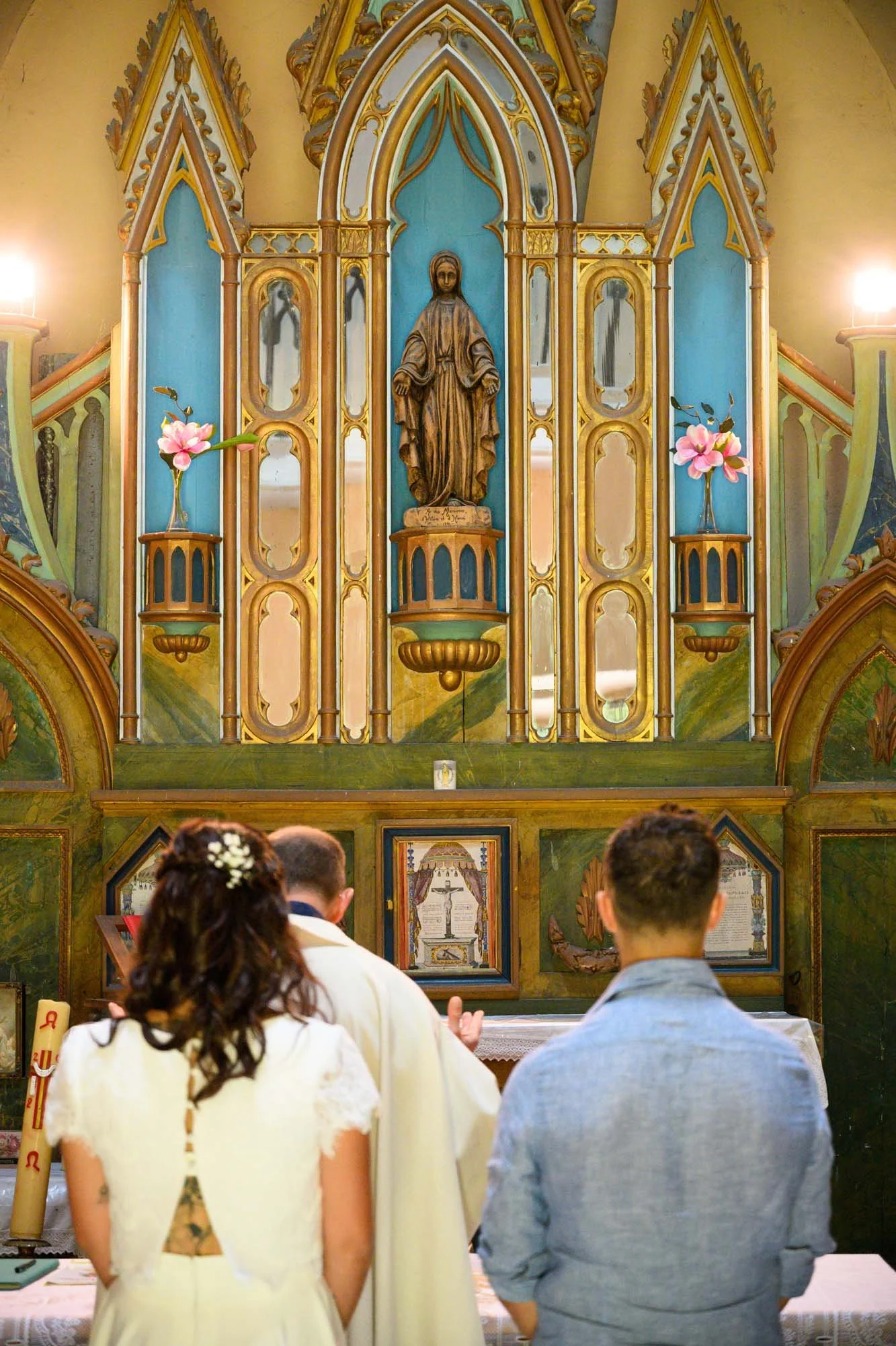 Un couple et un prêtre lors d'une cérémonie religieuse dans une église, avec un autel orné d'une statue religieuse et des décorations florales.