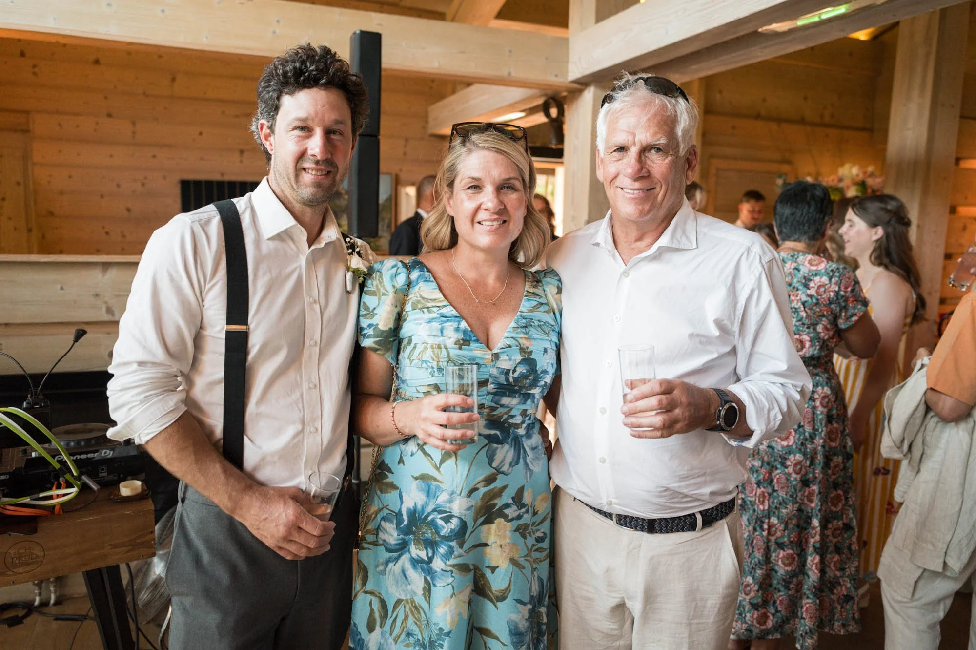 Trois personnes posant pour une photo lors d'une fête, deux hommes et une femme tenant des verres. L'ambiance est chaleureuse avec d'autres invités en arrière-plan.
