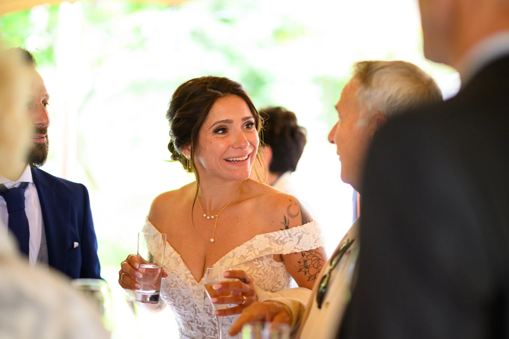Une femme en robe de mariée sourit en conversant avec un homme lors d'un mariage, tenant un verre dans chaque main.