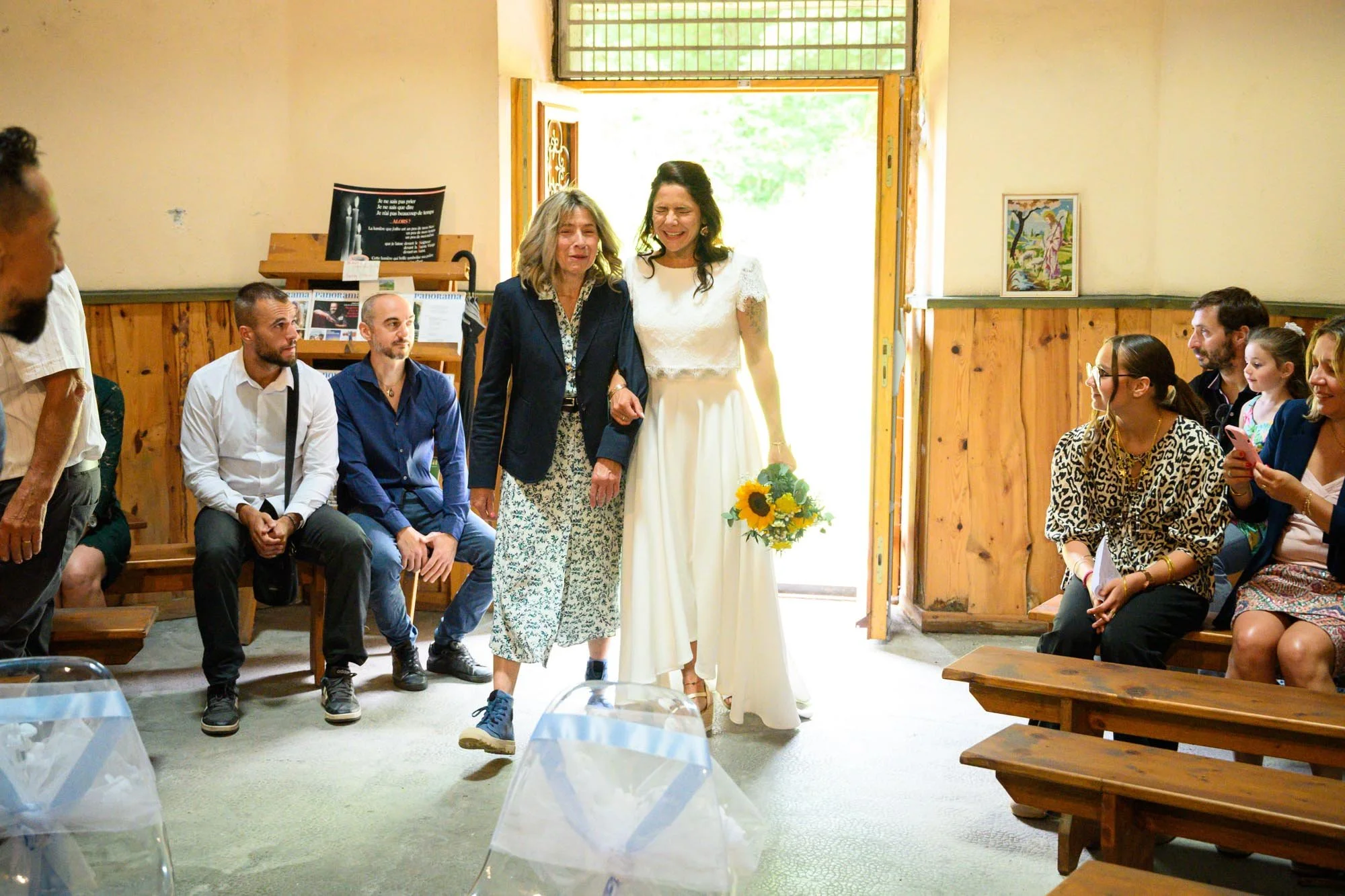 Une mariée en robe blanche tenant un bouquet de tournesols marche dans une pièce lors d'un mariage, accompagnée d'une femme plus âgée vêtue d'un blazer noir et d'une robe à motif. Des invités assis sur des bancs, certains prenant des photos, assisten