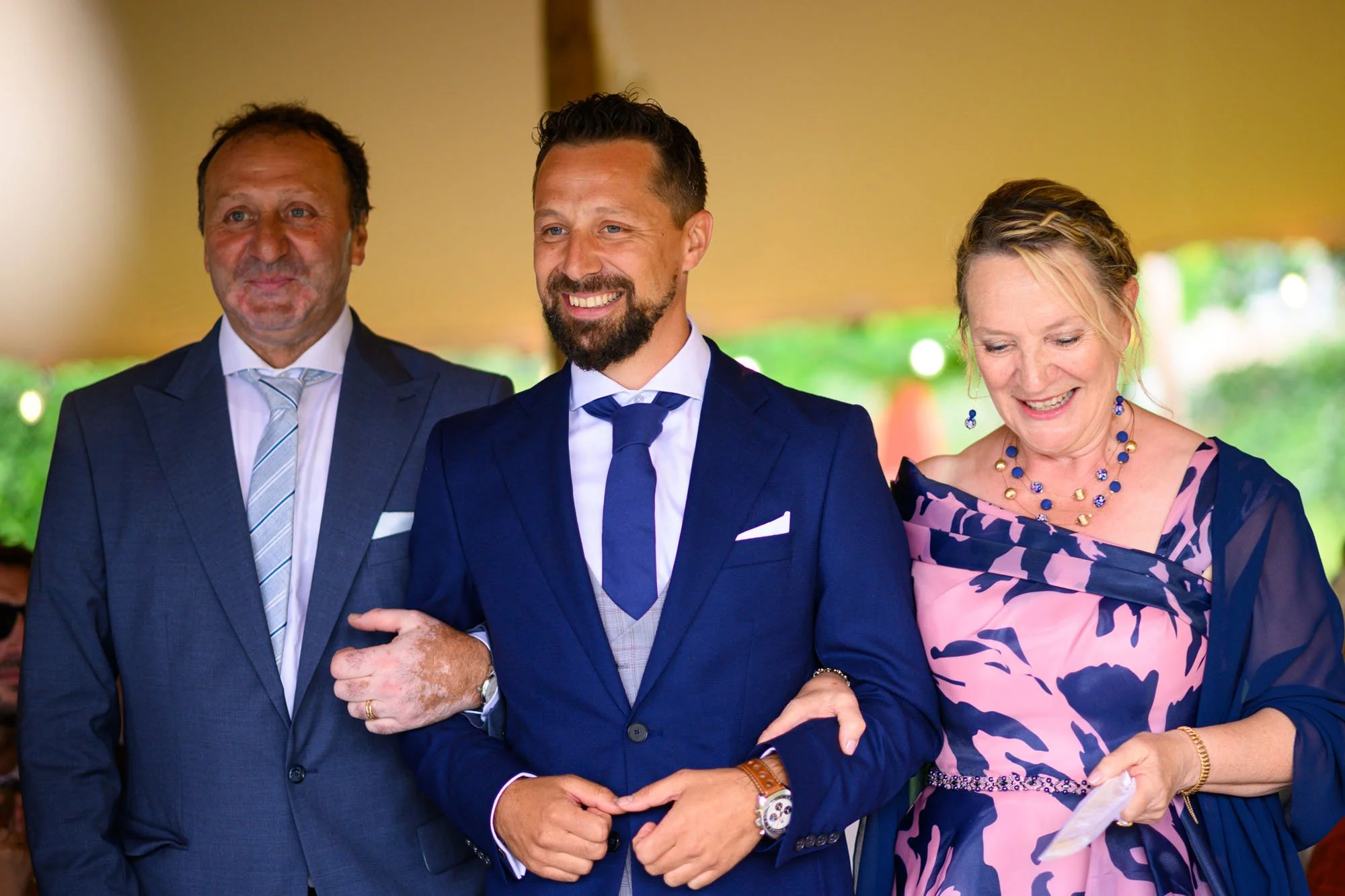 Trois personnes habillées en costume sont souriantes lors d'une cérémonie, une femme en robe rose et bleu, deux hommes en costume bleu.