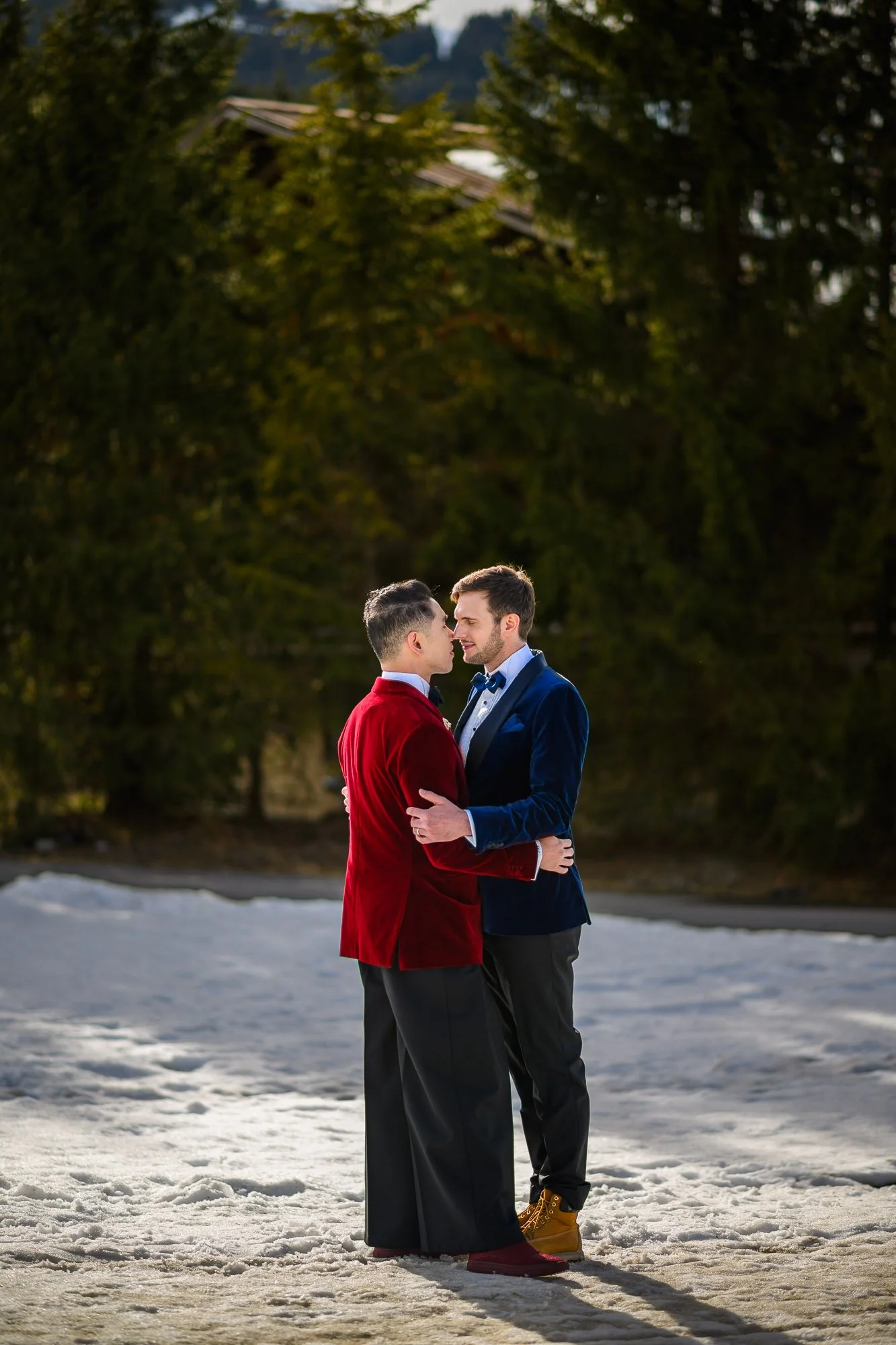 Deux hommes élégamment habillés, se tenant face à face sur la neige, partageant un moment intime en extérieur avec un fond de forêt et de montagnes.