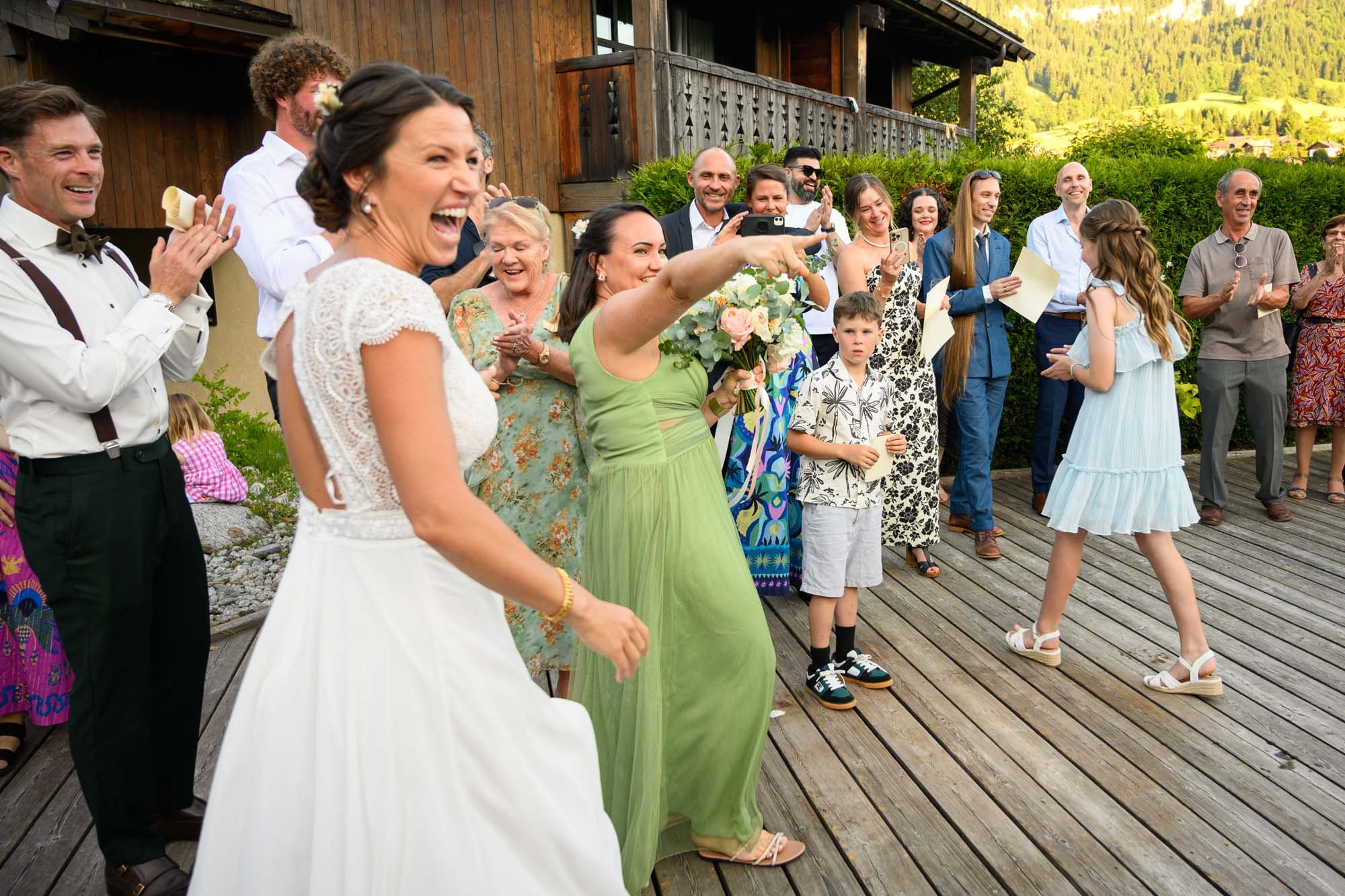 Groupe de personnes lors d'une célébration en plein air sur une terrasse en bois, avec une femme en robe blanche au premier plan, souriant et une autre femme en vert pointant, entourées d'adolescents et d'adultes, avec un paysage verdoyant en arrière