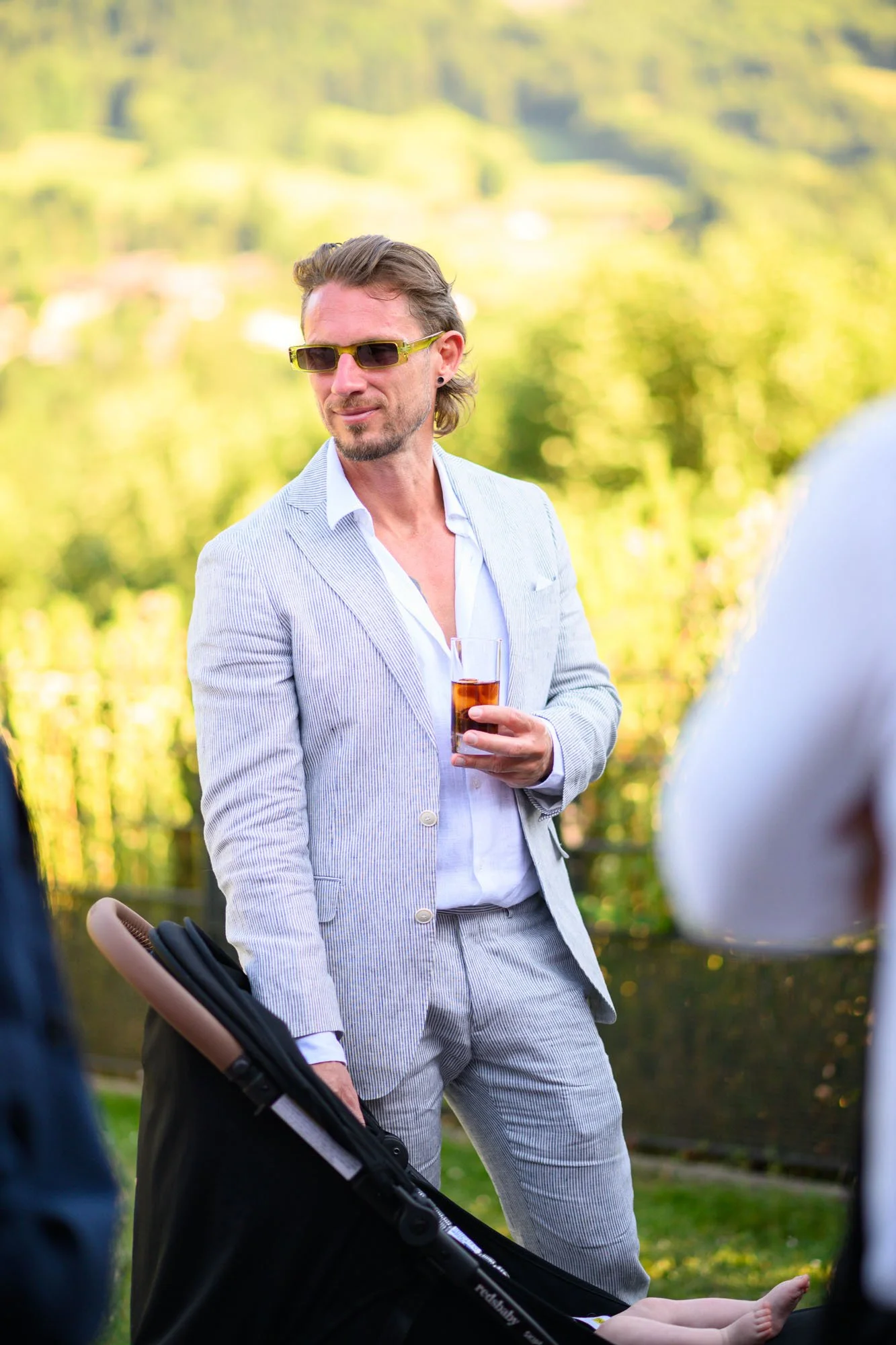 Un homme portant un costume rayé clair, sans cravate, avec des lunettes de soleil et une barbe, tient un verre de boisson dans une main, lors d'un événement en extérieur avec un fond de nature verdoyante.