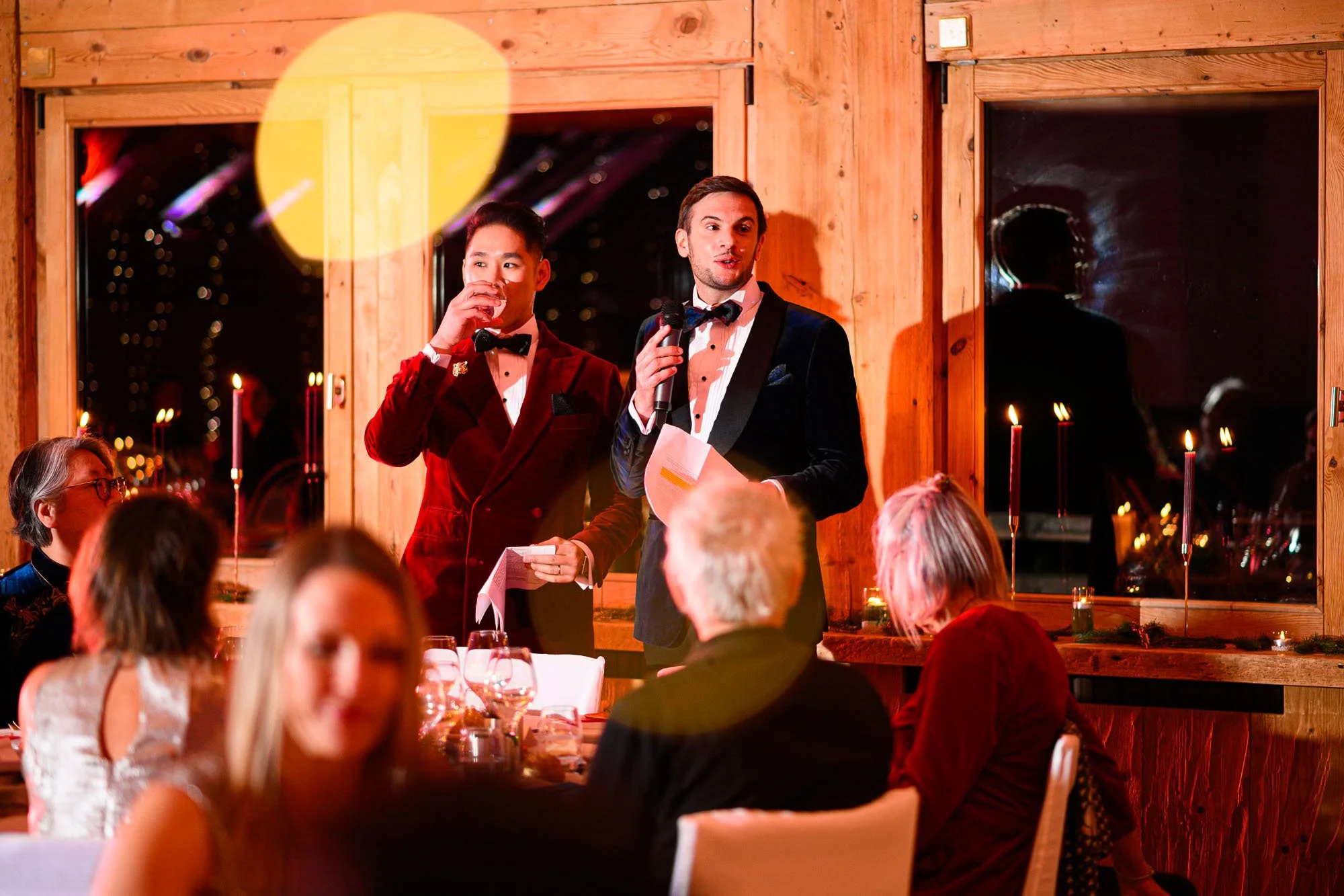 Deux hommes en costume tuxedo parlent lors d'un dîner formel dans une salle en bois avec des fenêtres, un public écoute.