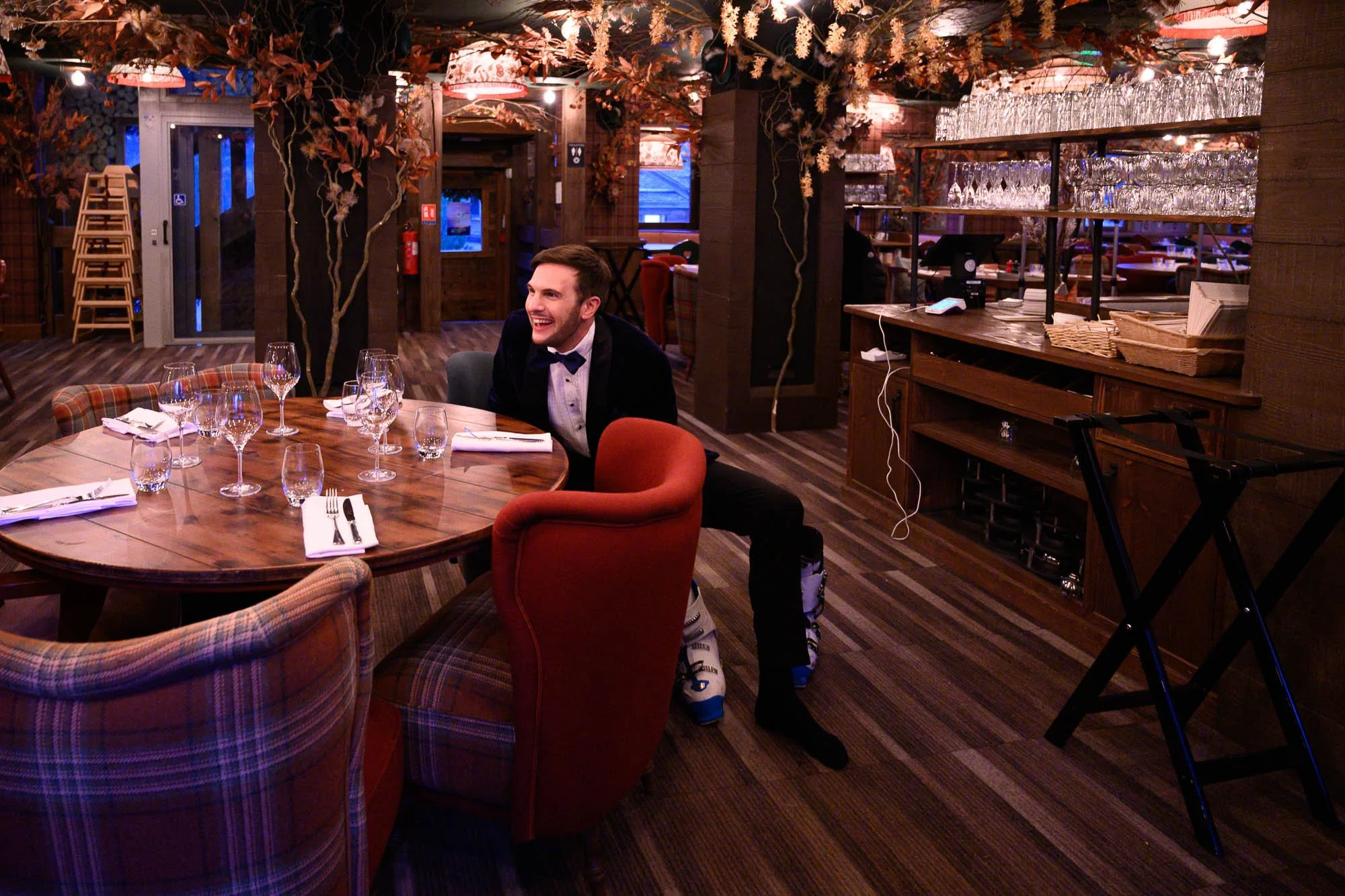 Un homme en smoking assis à une table de restaurant élégamment décorée, souriant, dans un intérieur chaleureux avec des lumières tamisées et des décorations florales.