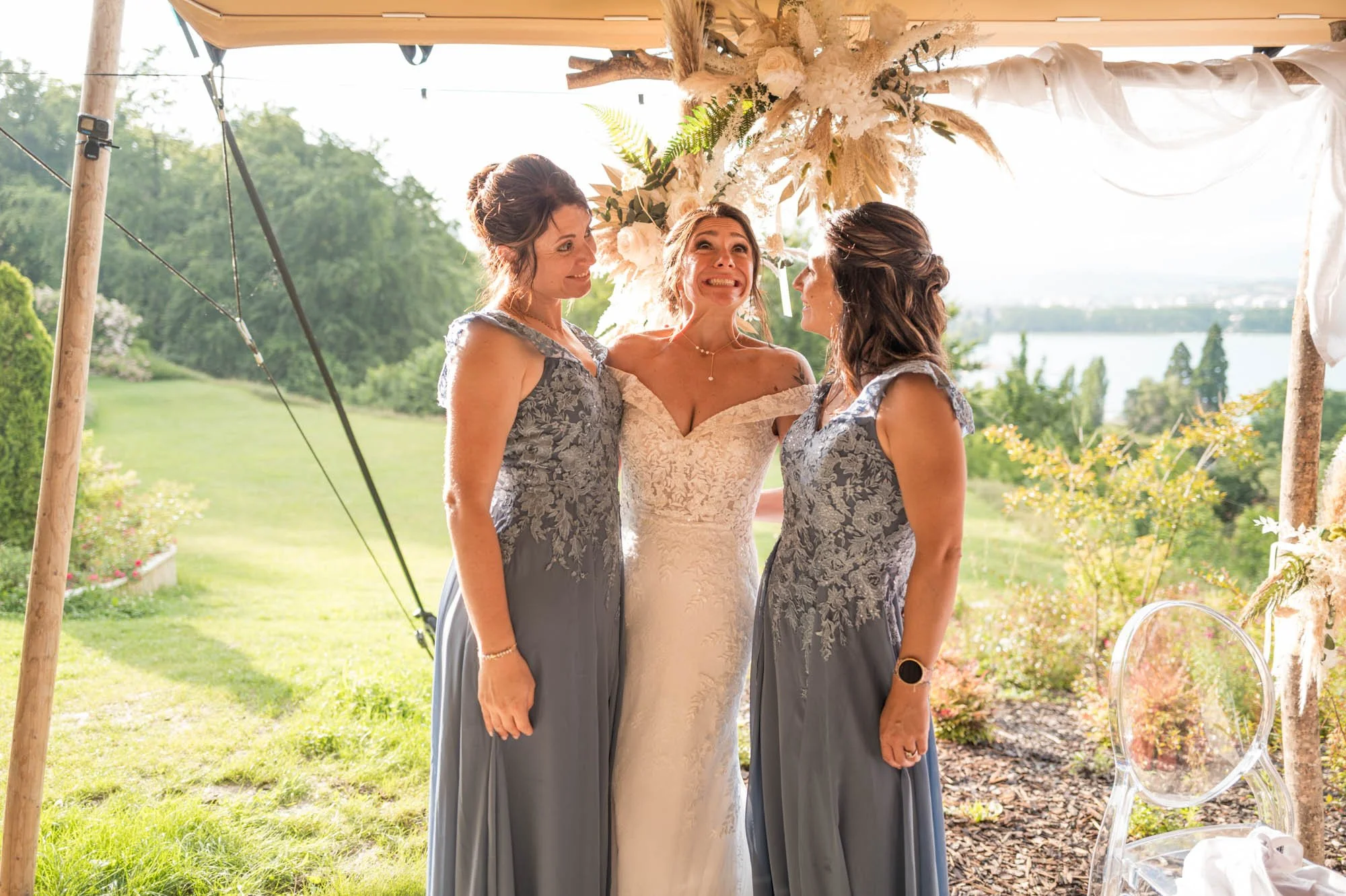 Une mariée souriante entourée de deux femmes en robes gris modernes, dans un décor extérieur lors d'un mariage. En arrière-plan, un paysage verdoyant et un lac.