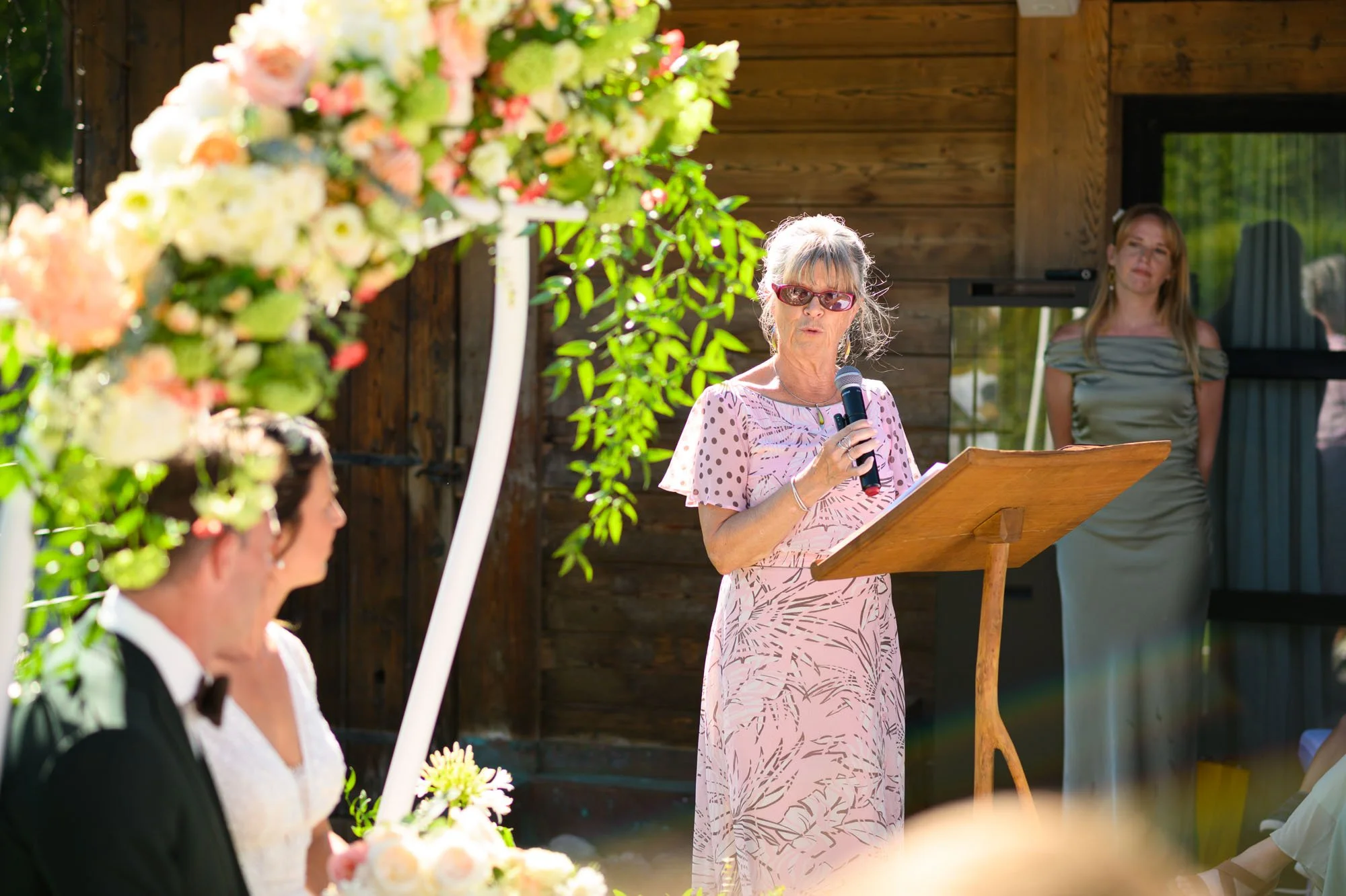 Une femme âgée parle lors d'un mariage en extérieur, devant une floral arrangement.