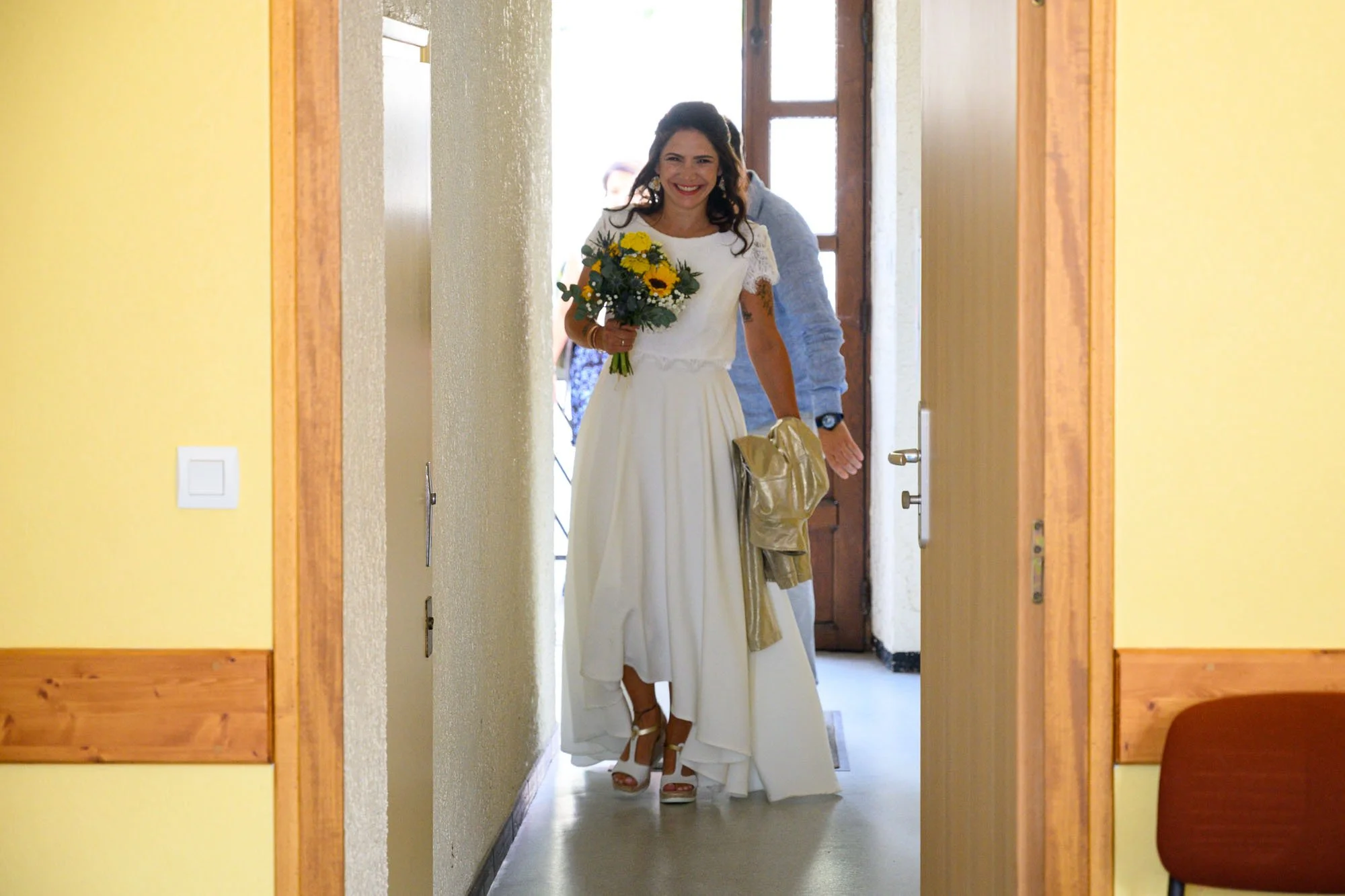 Une femme souriante en robe de mariée blanche, tenant un bouquet de fleurs, entre dans une pièce, accompagnée d'une personne hors champ.