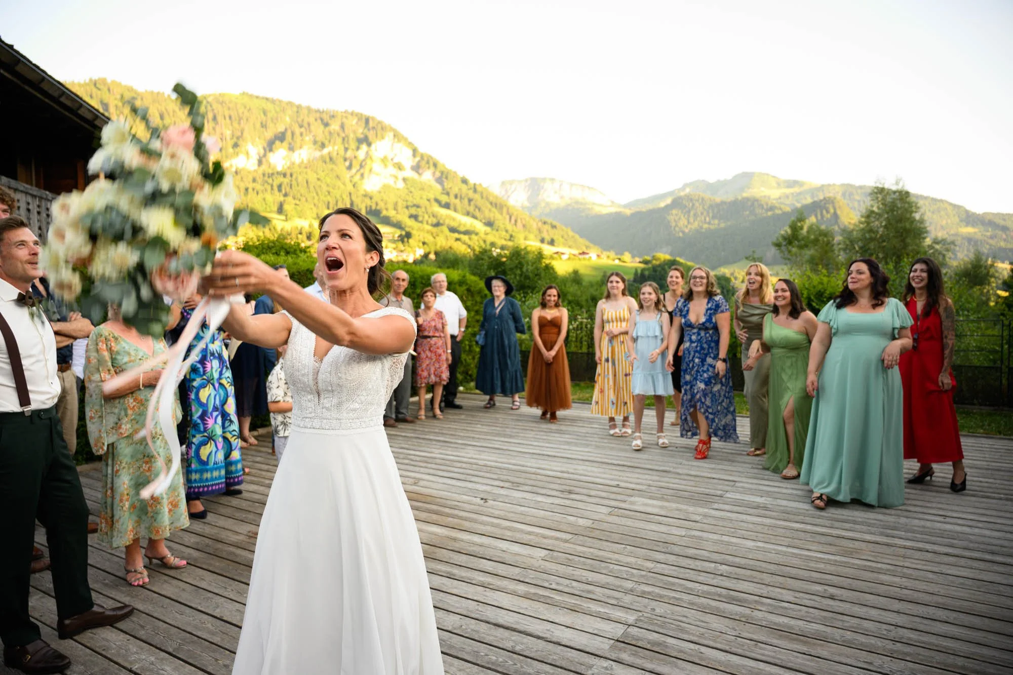 Une mariée en robe blanche lance un bouquet de fleurs aux invités lors d'un mariage en plein air, avec un groupe de femmes souriantes en arrière-plan et un paysage montagneux.