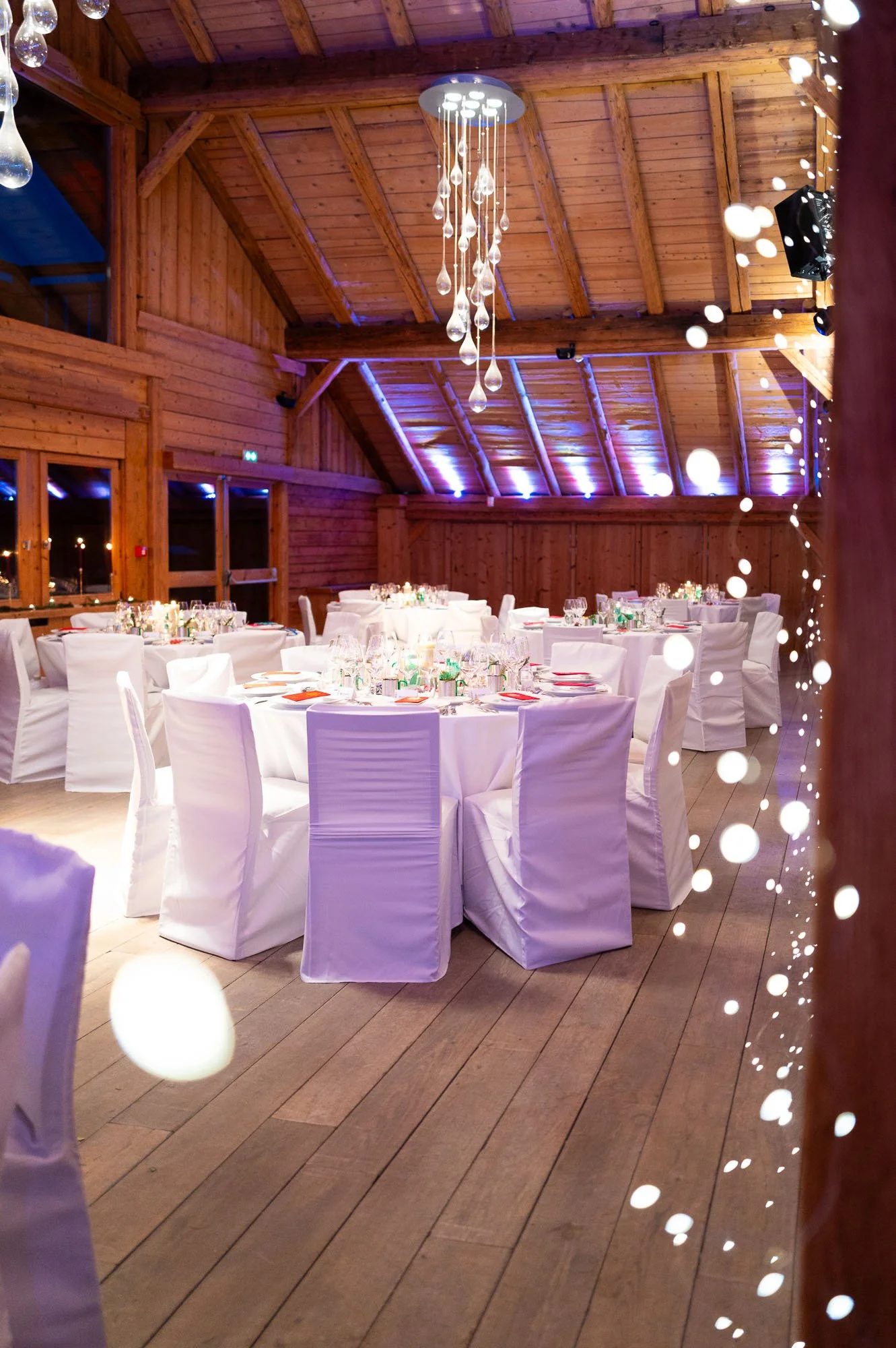 Salle de banquet en bois avec tables rondes, chaises habillées de blanc, décorations élégantes, éclairage tamisé, chandelier suspendu, ambiance festive pour un événement spécial.