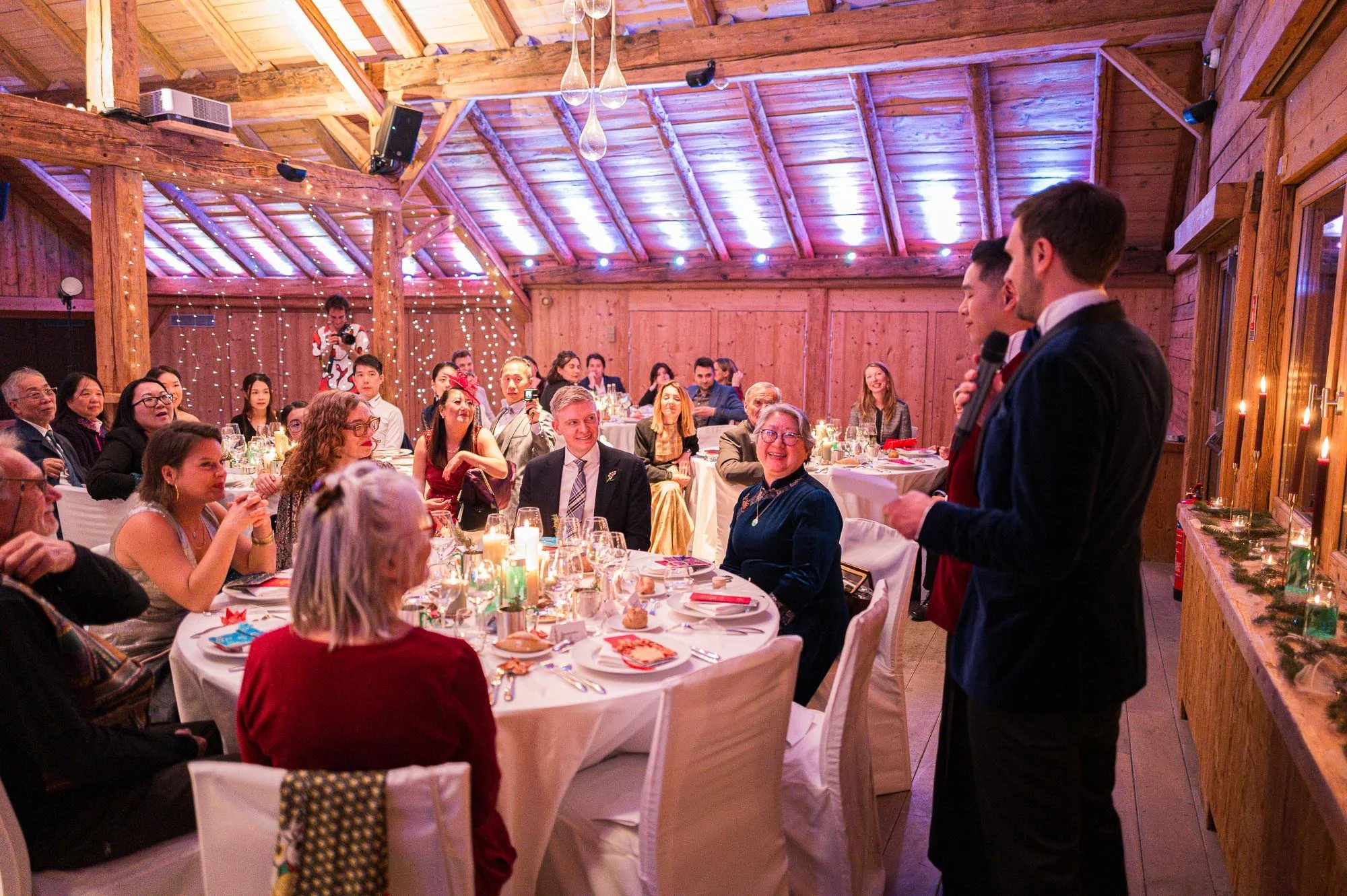 Un groupe de personnes assises autour d'une table lors d'une fête ou d'un dîner dans une salle en bois, avec deux hommes en costume noir donnant un discours ou une toast devant l'assemblée.