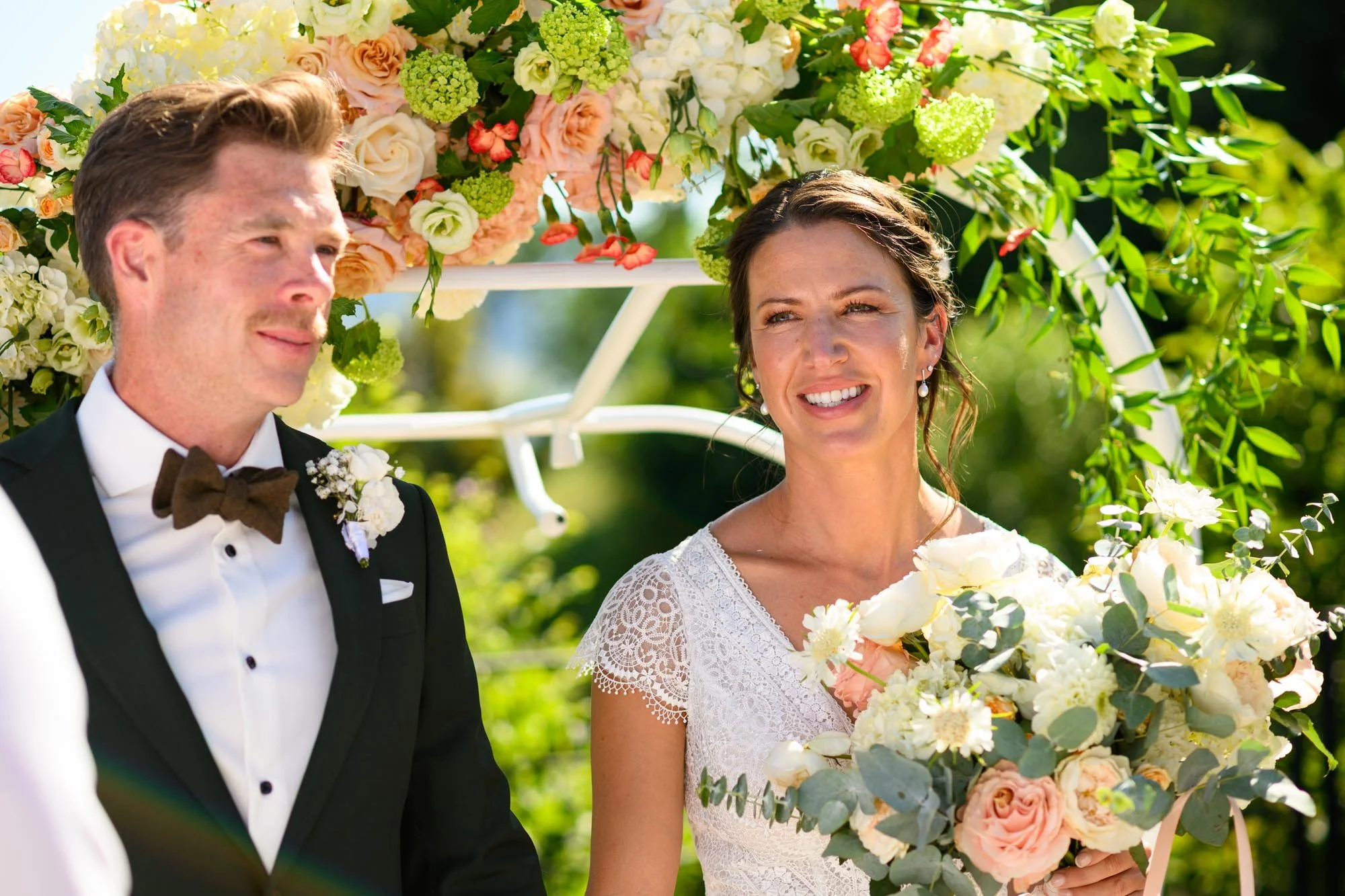 Un couple en costume de mariage, la femme tenant un bouquet de fleurs, sous une décoration florale lors d'une cérémonie en plein air.