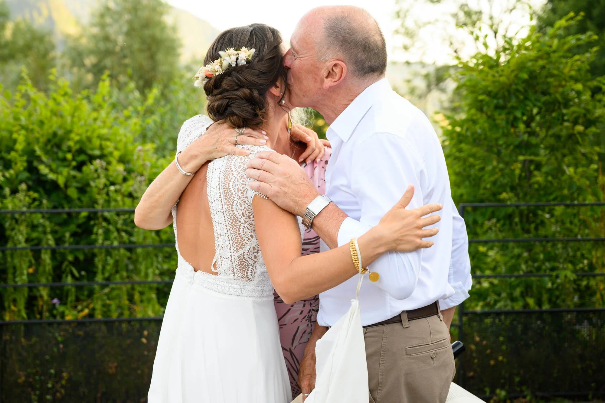 Un couple et une femme qui les entoure s'embrassent lors d'une célébration en plein air, probablement un mariage, avec des arbres verts en arrière-plan.