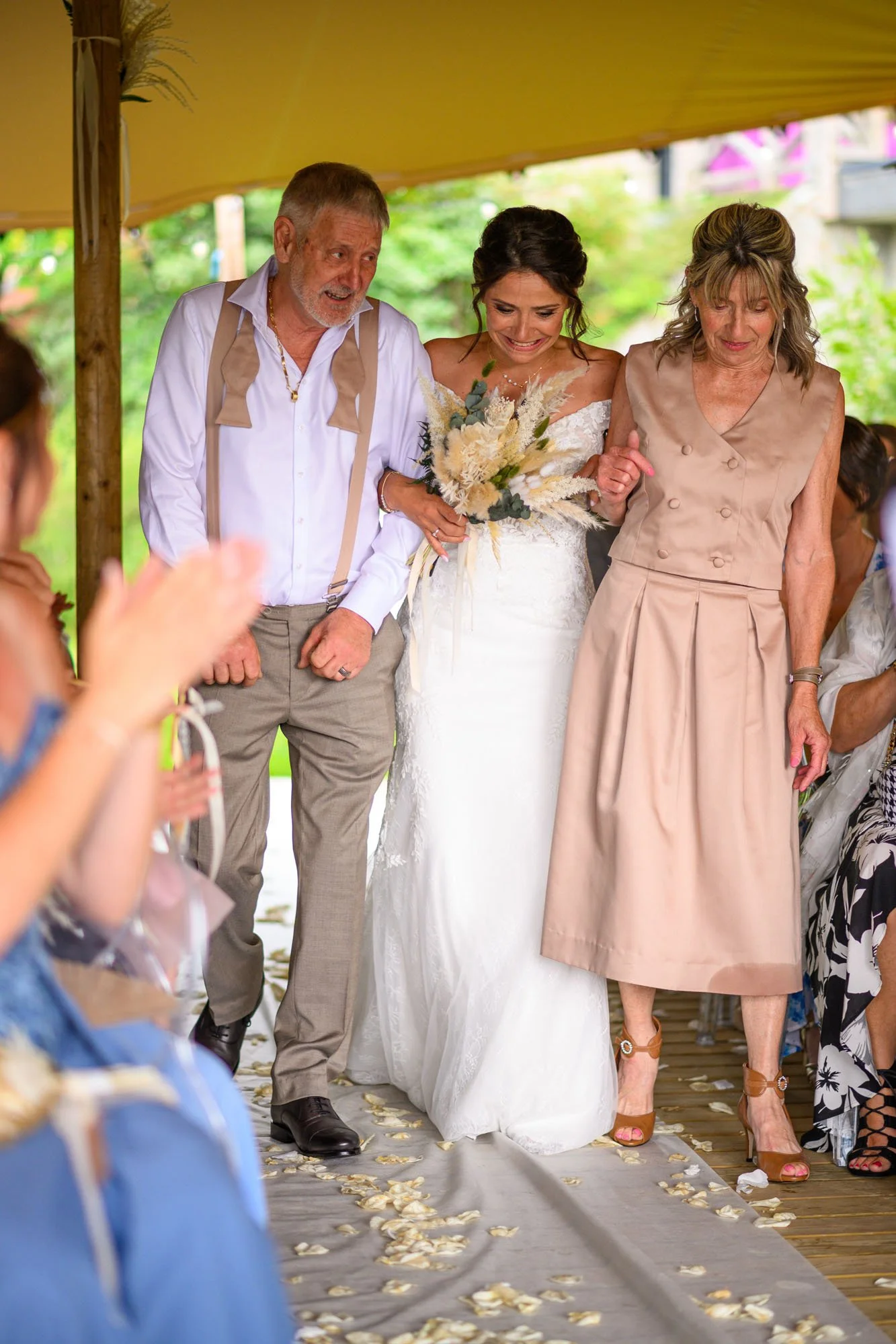 Une mariée en robe blanche marche dans une cérémonie de mariage, soutenue par deux personnes, entourée d'invités lors d'une célébration en plein air sous un abri en bois, avec des pétales de fleurs sur le sol.
