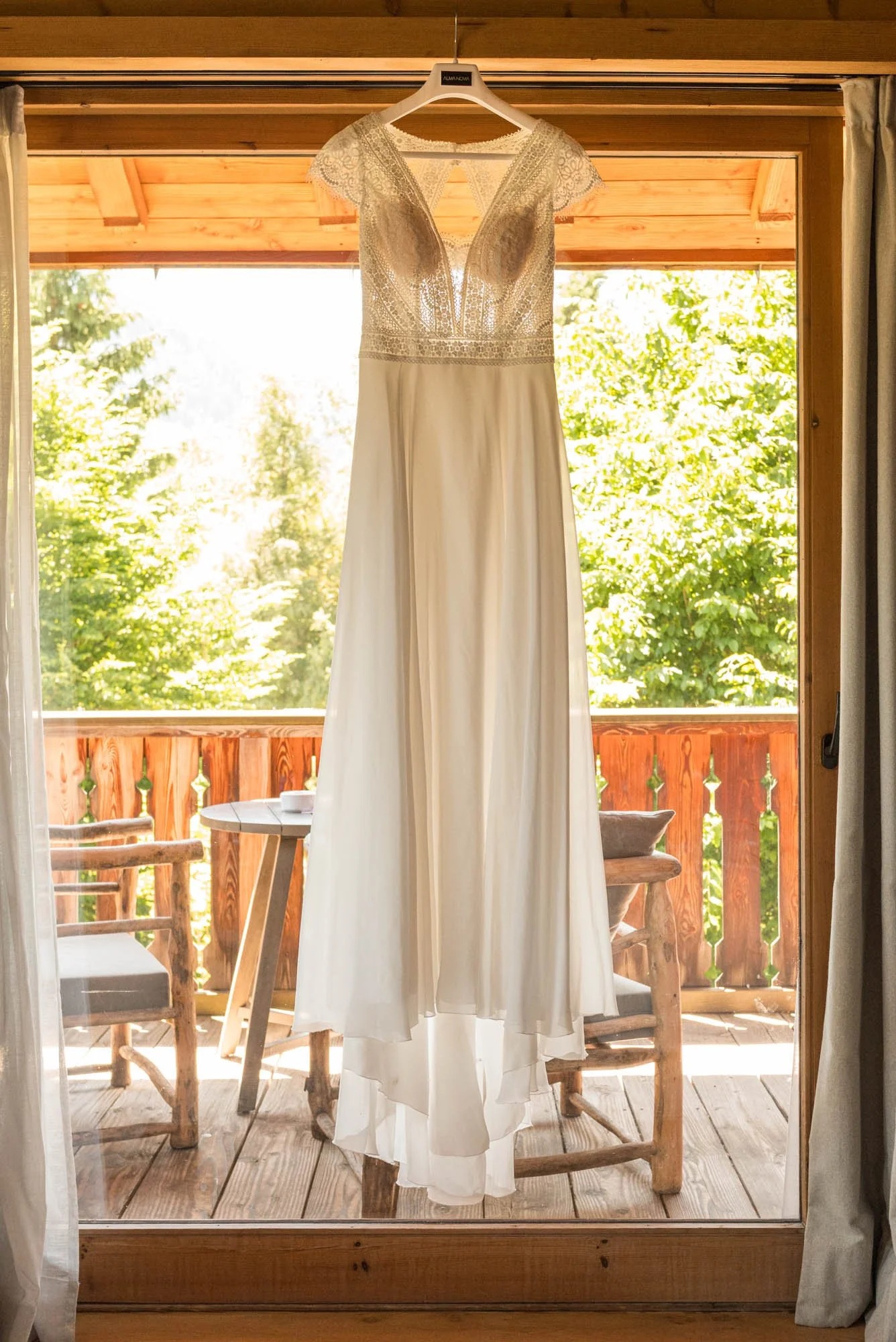 Robe de mariée blanche suspendue à une poignée de porte en bois avec un cadre en bois, vue d'une terrasse en bois avec des chaises en bois et une table, entourée de feuxillage vert et de ciel clair.