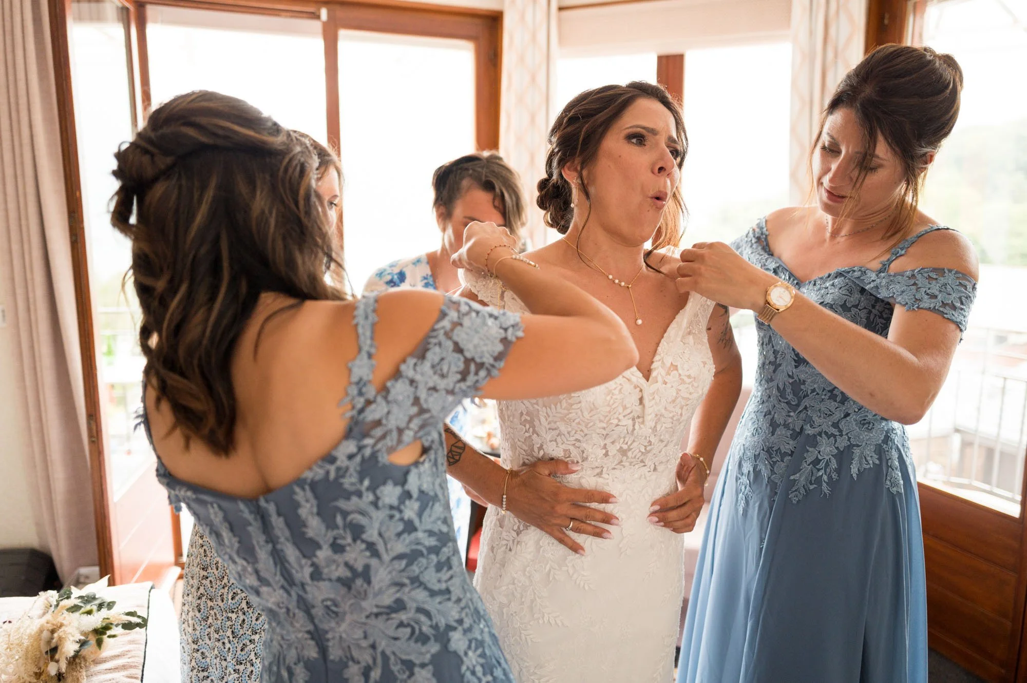 Une femme en robe de mariée est aidée par ses amies à se préparer dans une pièce lumineuse.