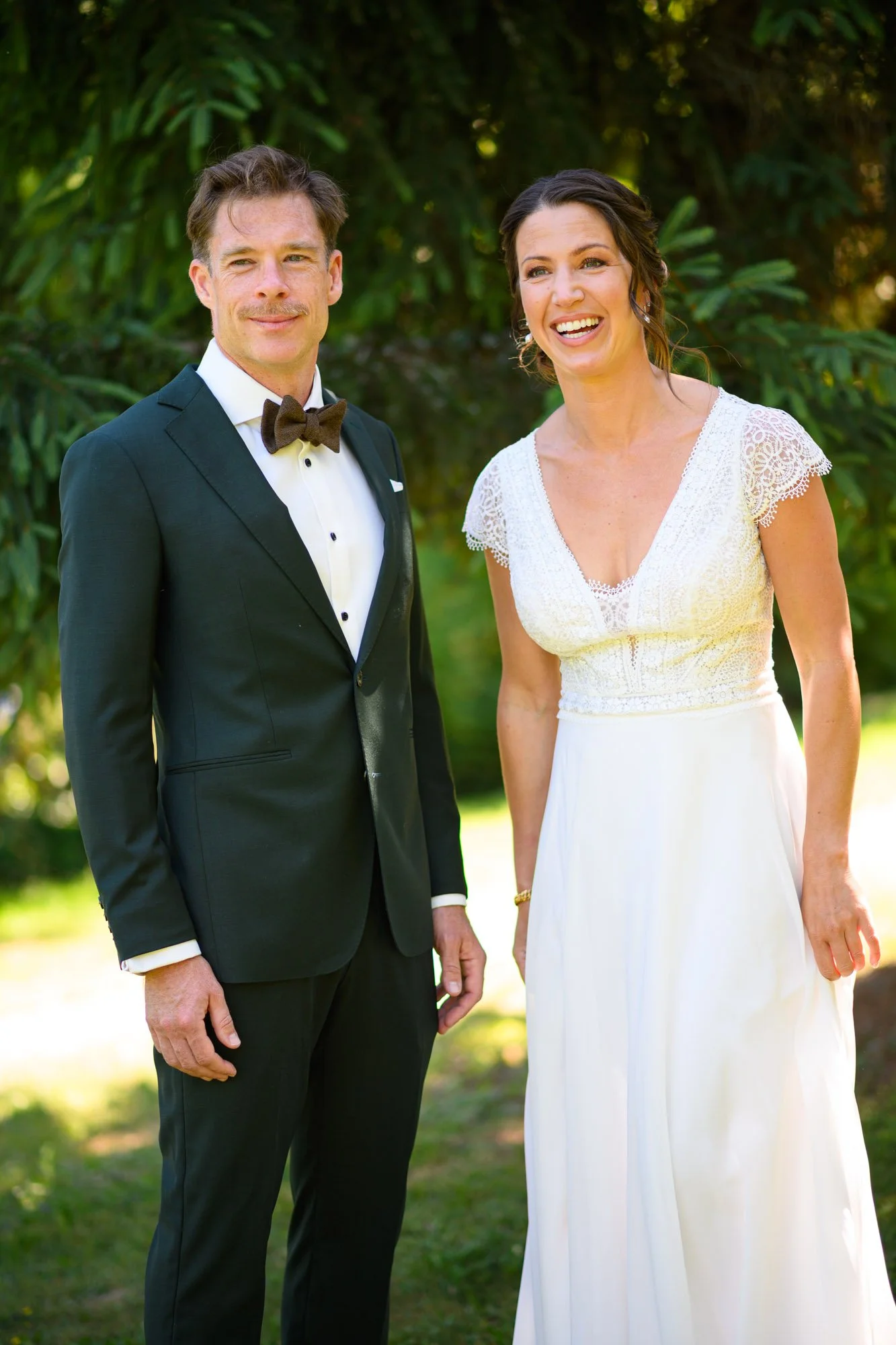 Un homme et une femme en tenue de mariage posent dehors devant des arbres, l'homme porte un smoking noir avec une chemise blanche et un nœud papillon, la femme porte une robe blanche en dentelle avec des manches courtes et un décolleté en V. La femme