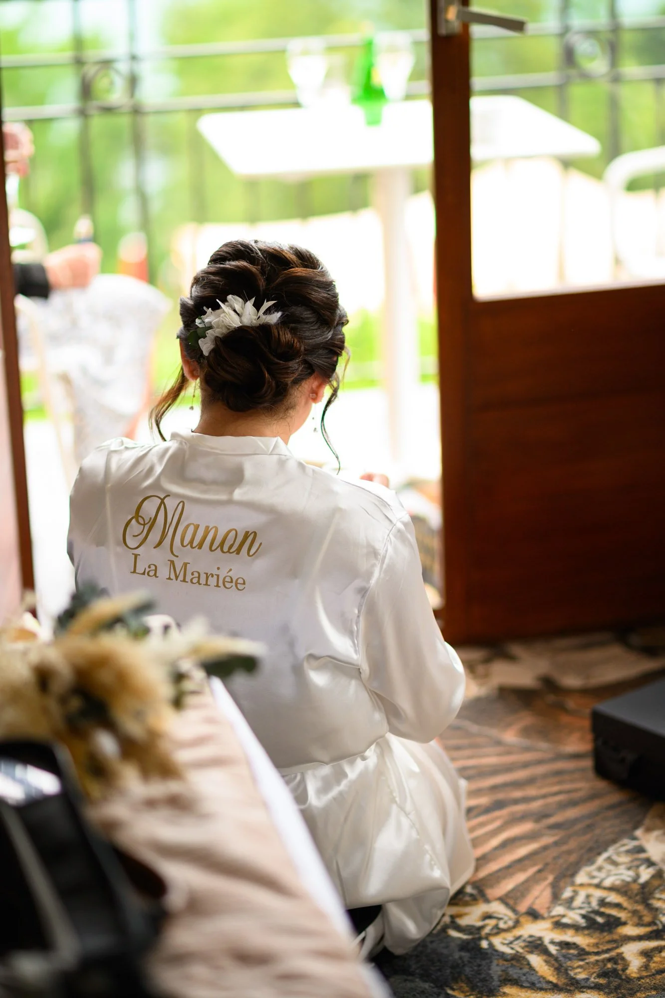 Une femme en robe de mariée vue de dos, portant une robe en satin blanc avec l'inscription 'Manan La Mariée' à l'arrière, assise à l'intérieur près d'une porte ouverte, avec un décor naturel en arrière-plan.