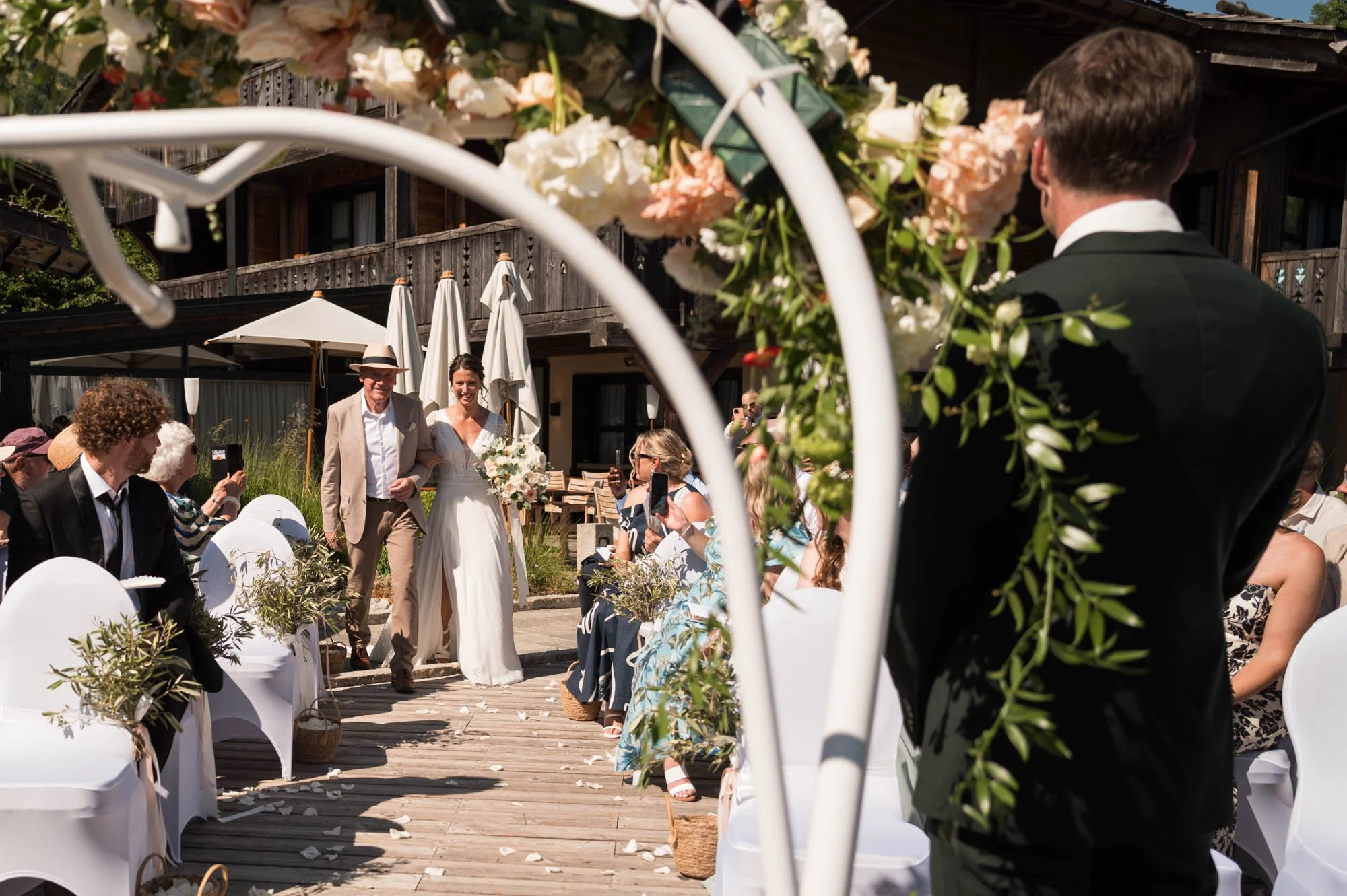Mariage en plein air avec la mariée marchant dans l'allée, entourée d'invités assis, décor floral, cadre en bois avec parasols blancs et parasol, ambiance ensoleillée.
