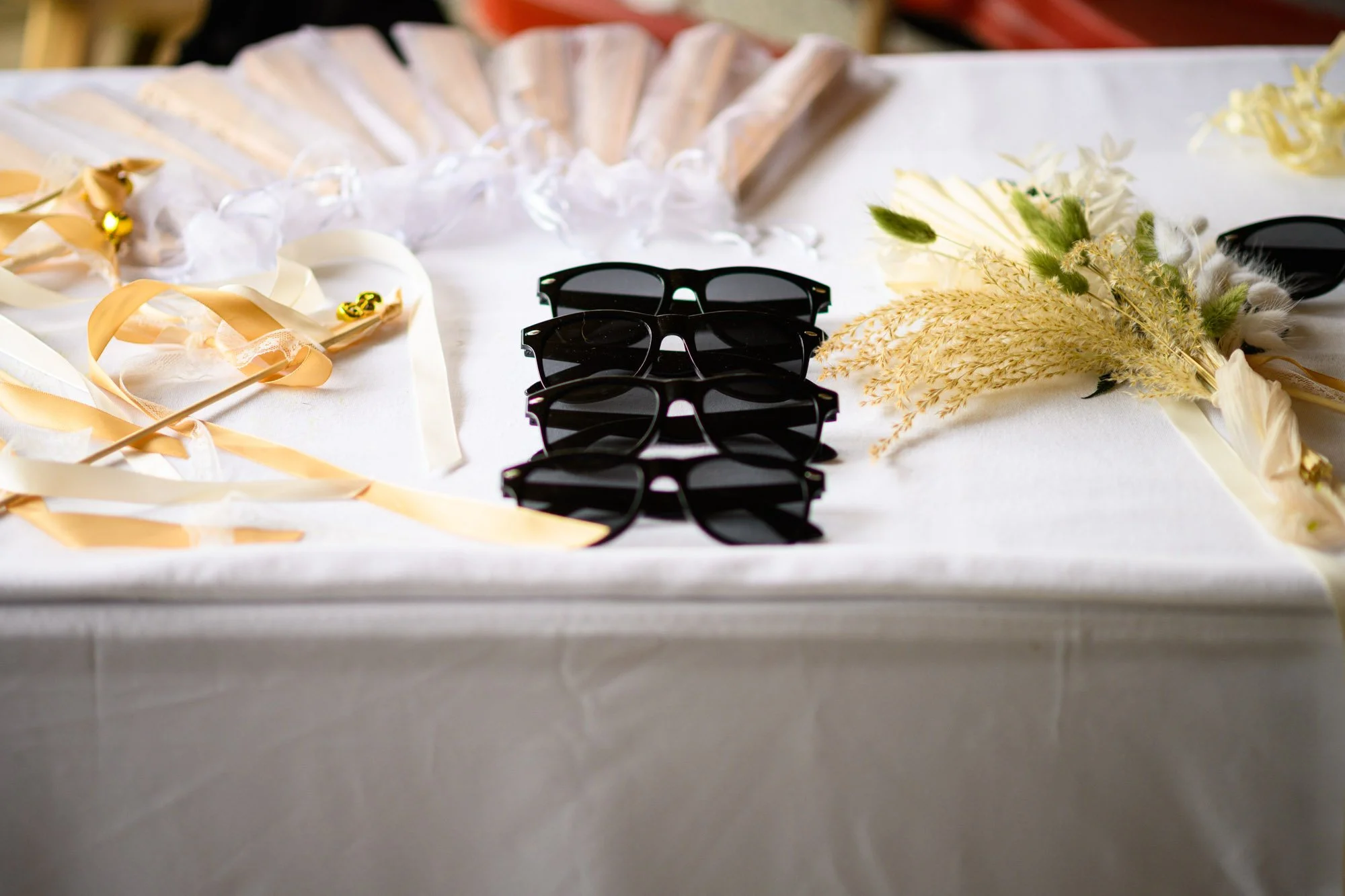 Quatre paires de lunettes de soleil noires placées en ligne sur une table recouverte d'une nappe blanche, entourées de rubans de satin beige, de plumes blanches et d'une petite branche de fleurs séchées, peut-être dans un cadre festif ou décoratif.