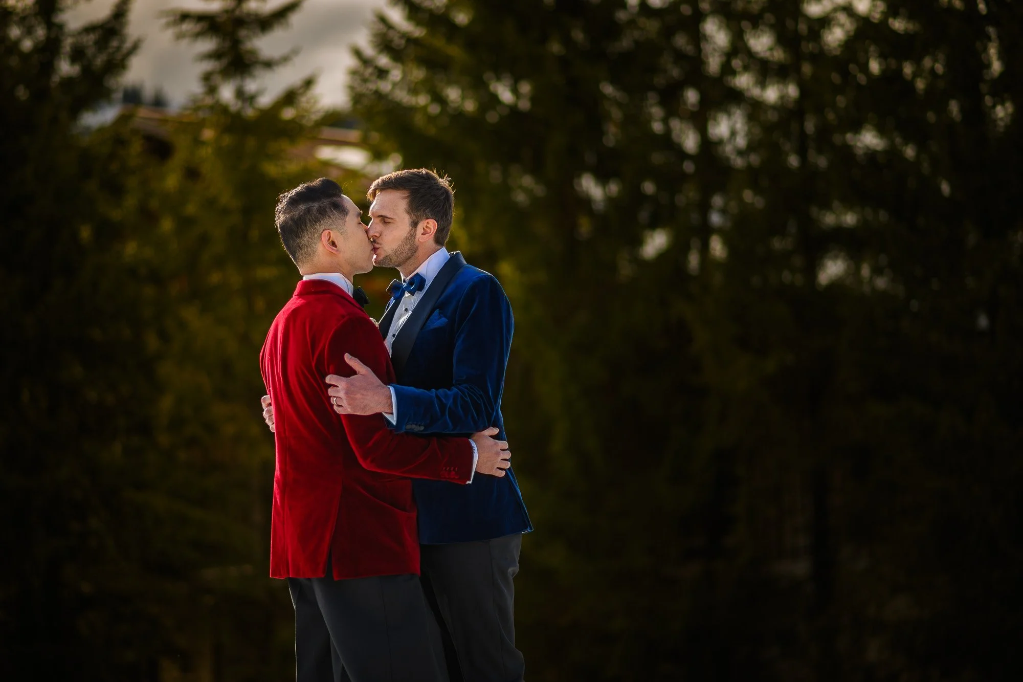 Deux hommes en costume se câlinent et s'embrassent dans un parc entouré d'arbres, lors d'une soirée.