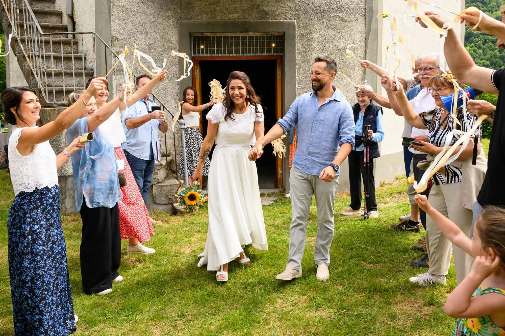 Un groupe de personnes lors d'une fête ou d'un mariage, formant une allée pour le couple central, avec des gens qui tiennent des rubans et s'amusent dehors devant un bâtiment.
