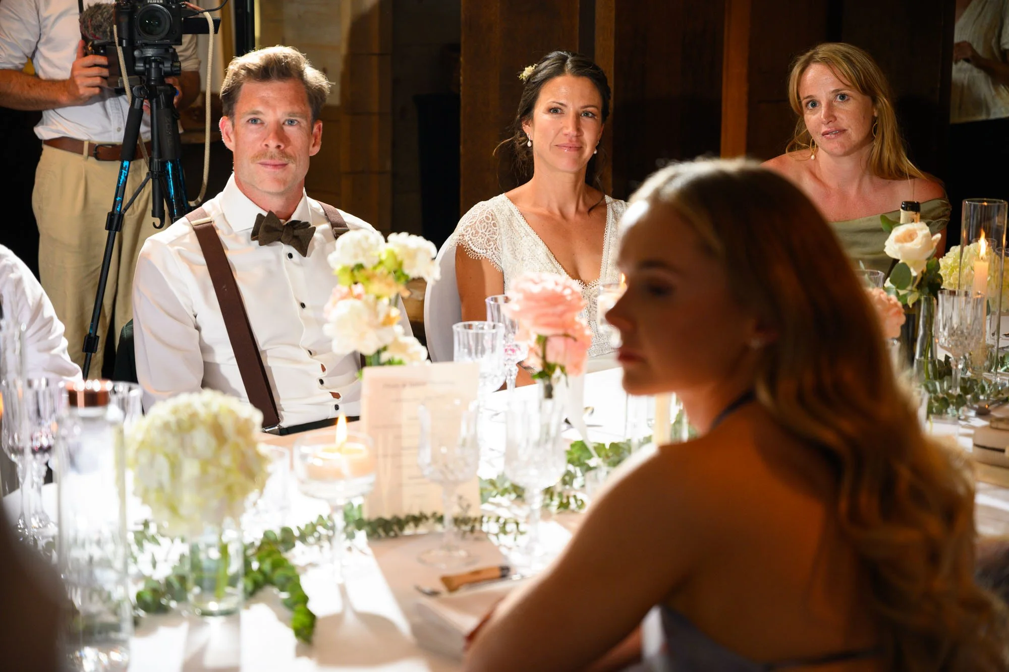 Gens assis à une table lors d'un mariage, décorée de fleurs et de bougies, avec un personnel de tournage en arrière-plan.