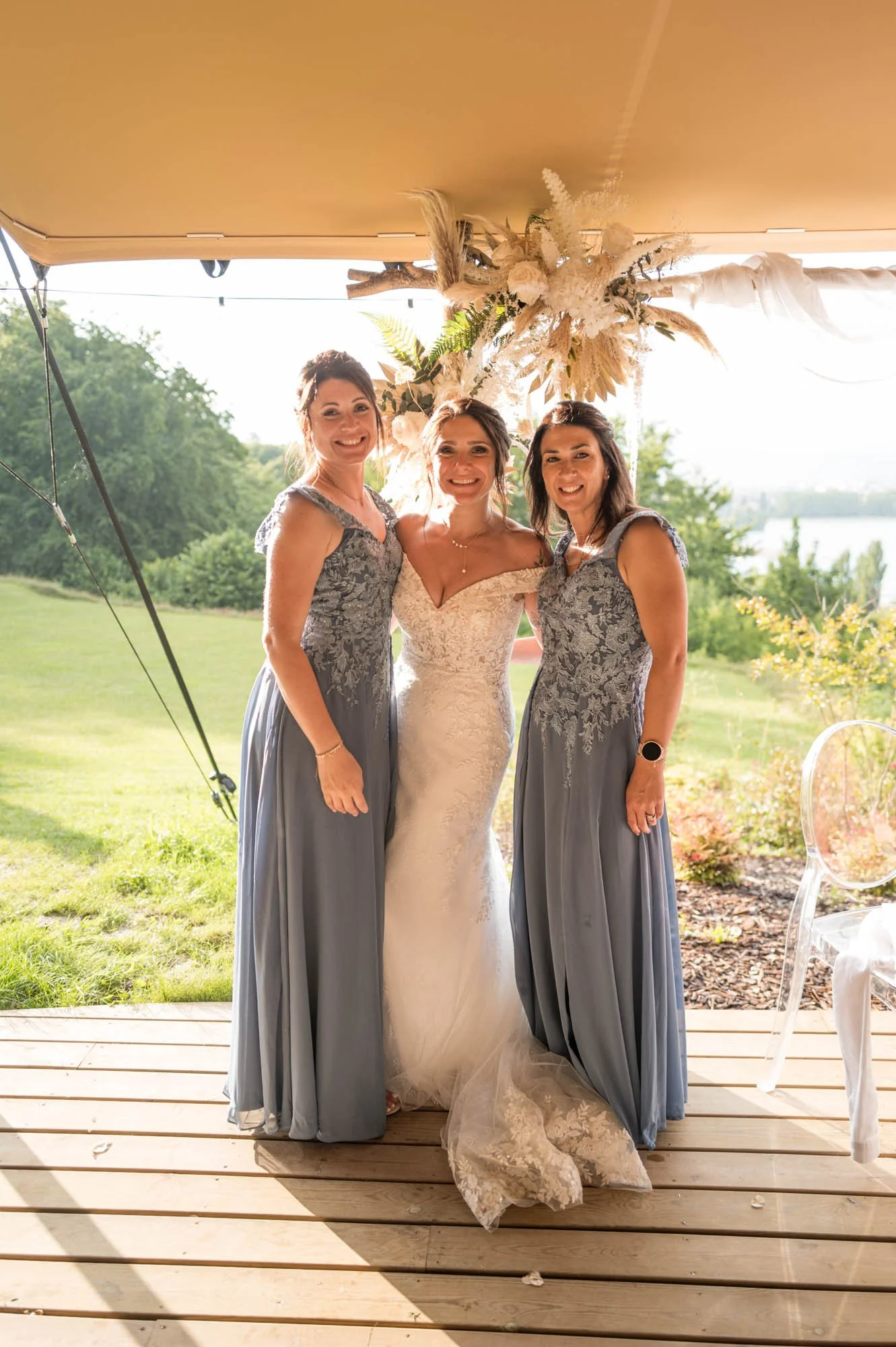 Trois femmes souriantes posant pour une photo lors d'un mariage en plein air, avec un décor floral blanc et vert en arrière-plan.