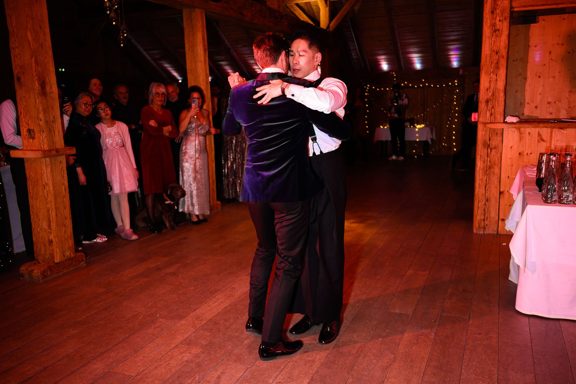 Deux hommes dansent ensemble dans une salle de réception, avec des invités qui regardent et prennent des photos en arrière-plan.