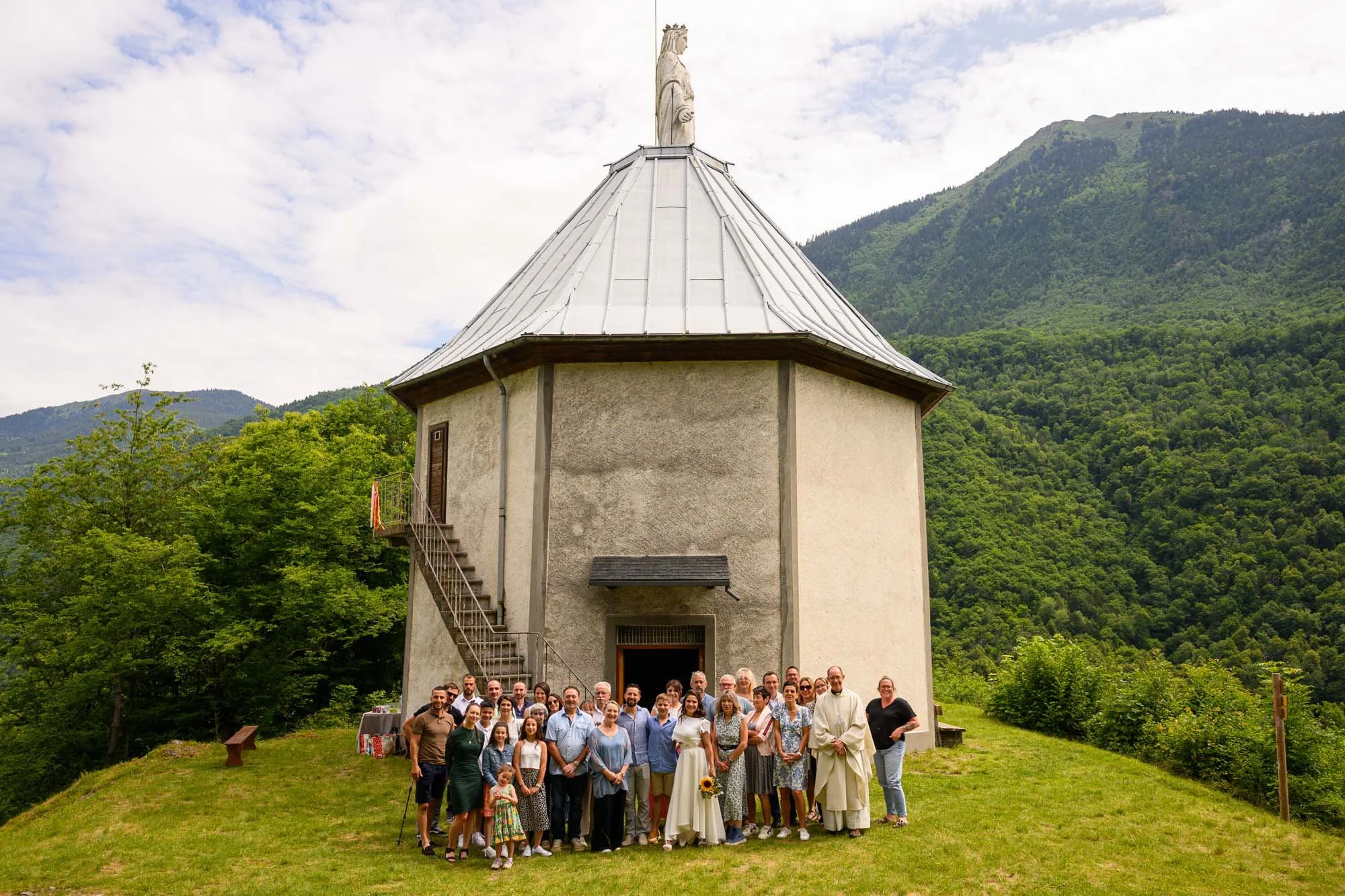 Groupe de personnes posant devant une petite église en pierre avec une statue religieuse au sommet, entourée de montagnes verdoyantes.
