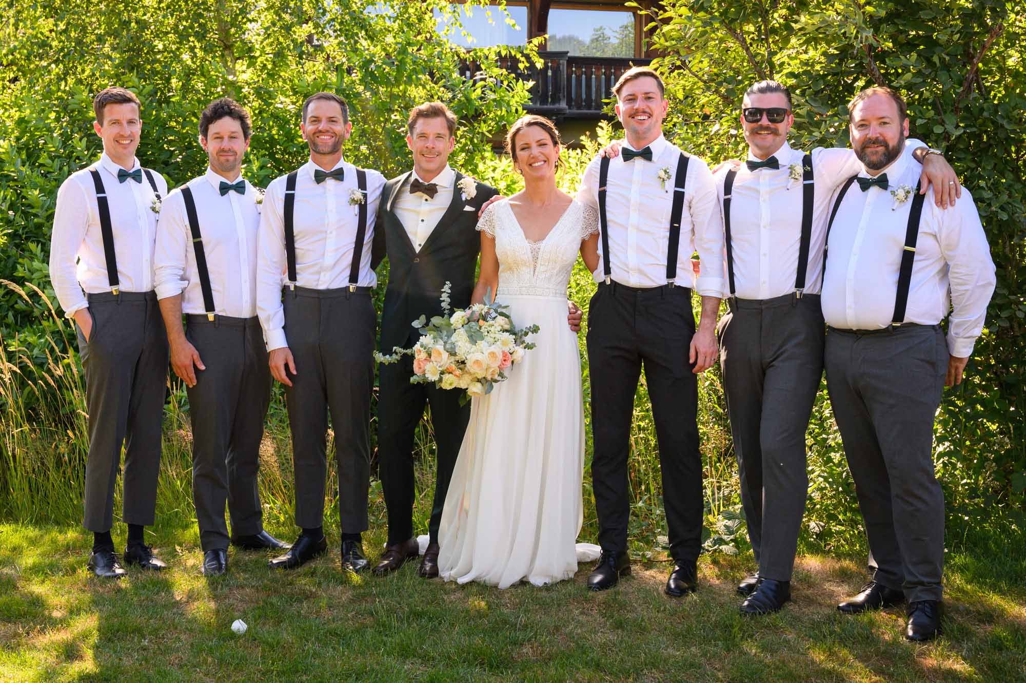 Groupe de huit personnes lors d'un mariage en extérieur, comprenant le marié, la mariée et six hommes en tenue formelle avec chemises blanches, pantalons gris et bretelles, dans un jardin avec des arbres et une maison en arrière-plan.