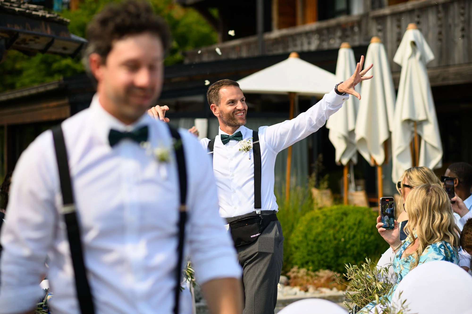 Un homme en costume blanc, chemise et nœud papillon, souriant et levant la main lors d'une célébration en plein air, entouré d'invités.