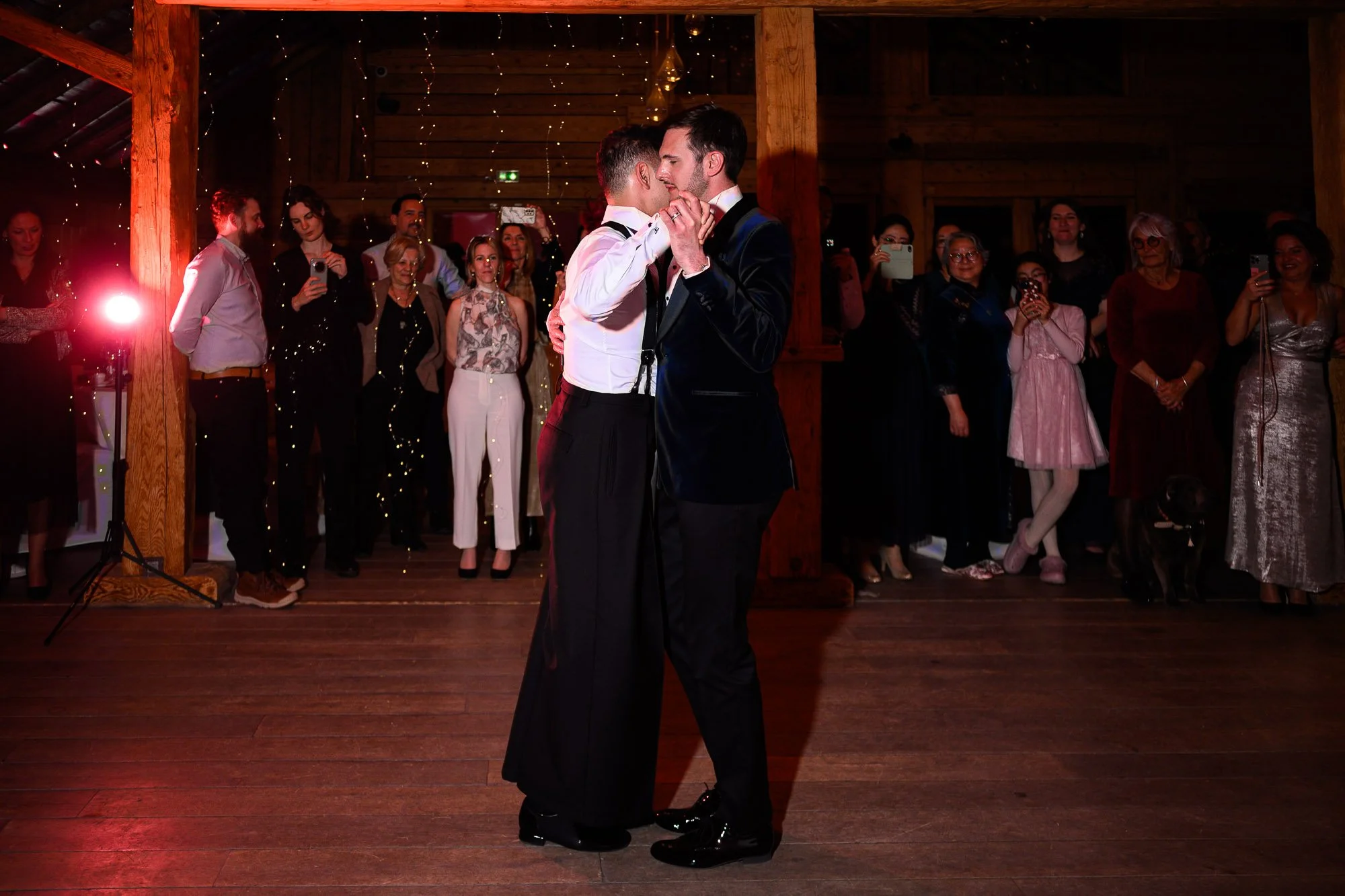 Deux hommes dansent et s'embrassent lors d'une surprise dans le cadre d'une cérémonie ou d'une célébration, avec un groupe de spectateurs en arrière-plan dans une salle en bois et décorée de guirlandes lumineuses.