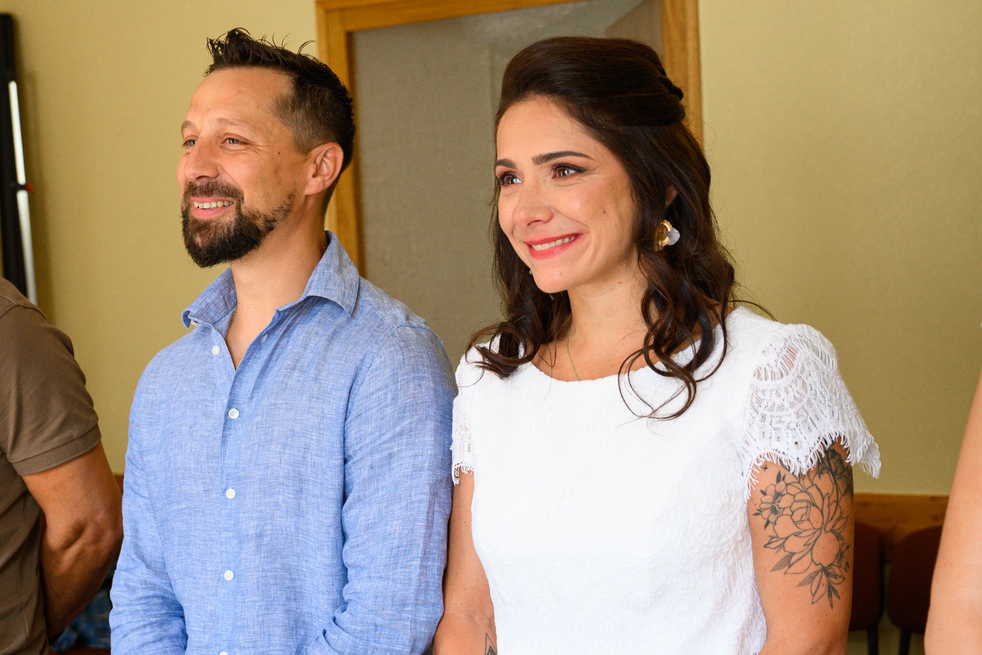 Deux personnes souriantes, un homme à gauche avec une barbe et une chemise bleue, et une femme à droite avec des cheveux bruns bouclés, une robe blanche avec des manches en dentelle, et un tatouage de fleurs sur le bras, posent dans une pièce.