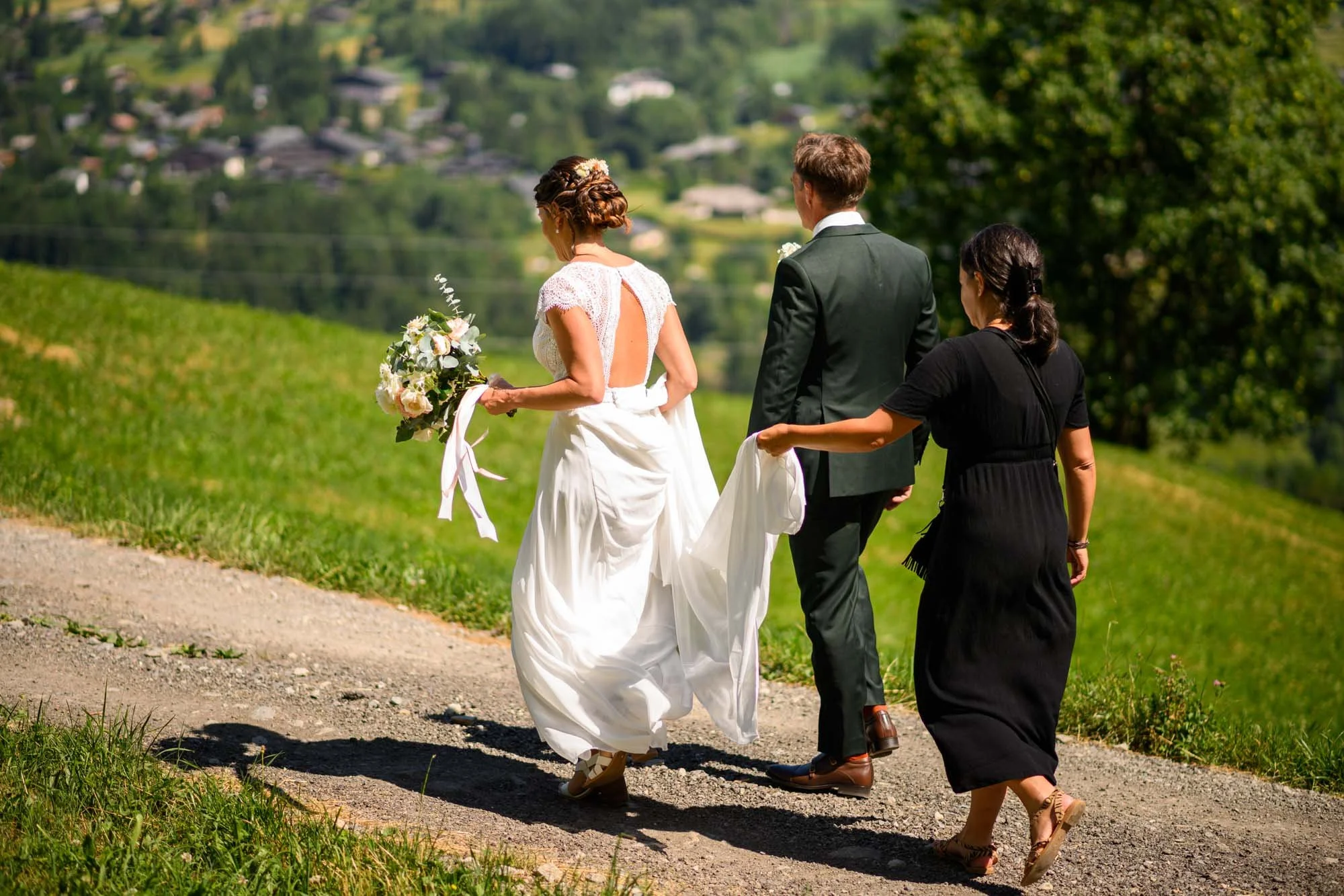 Un couple de mariés marche dans la nature avec une femme qui porte la robe de la mariée, sur un sentier en pleine campagne, entouré d'arbres et de verdure.