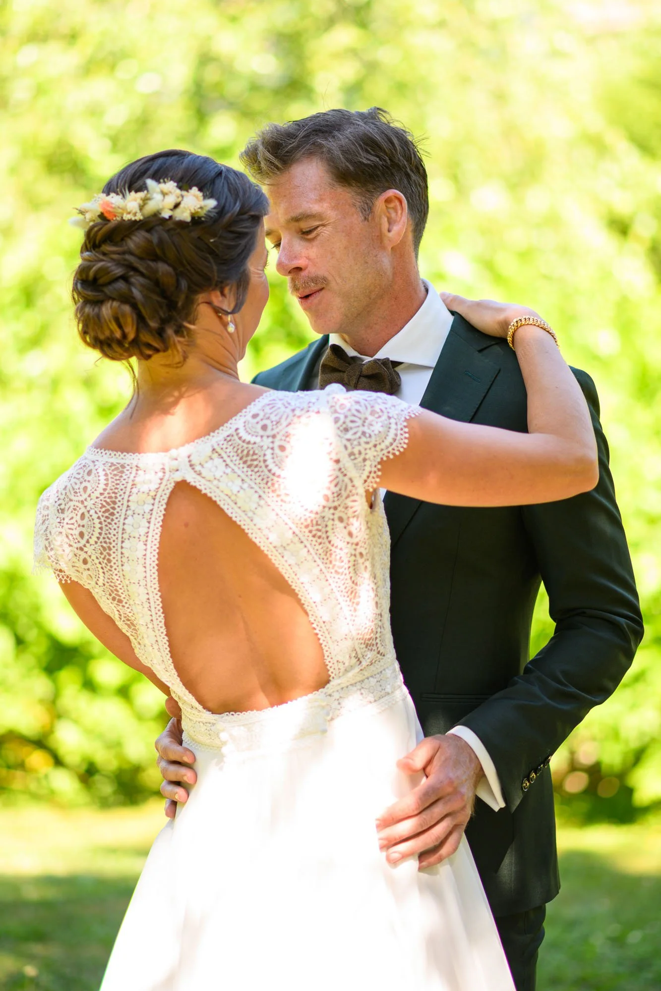 Un couple de mariés danse lors d'un mariage en plein air dans un jardin vert, avec un homme en costume noir et une femme en robe de mariée avec un dos nu en dentelle