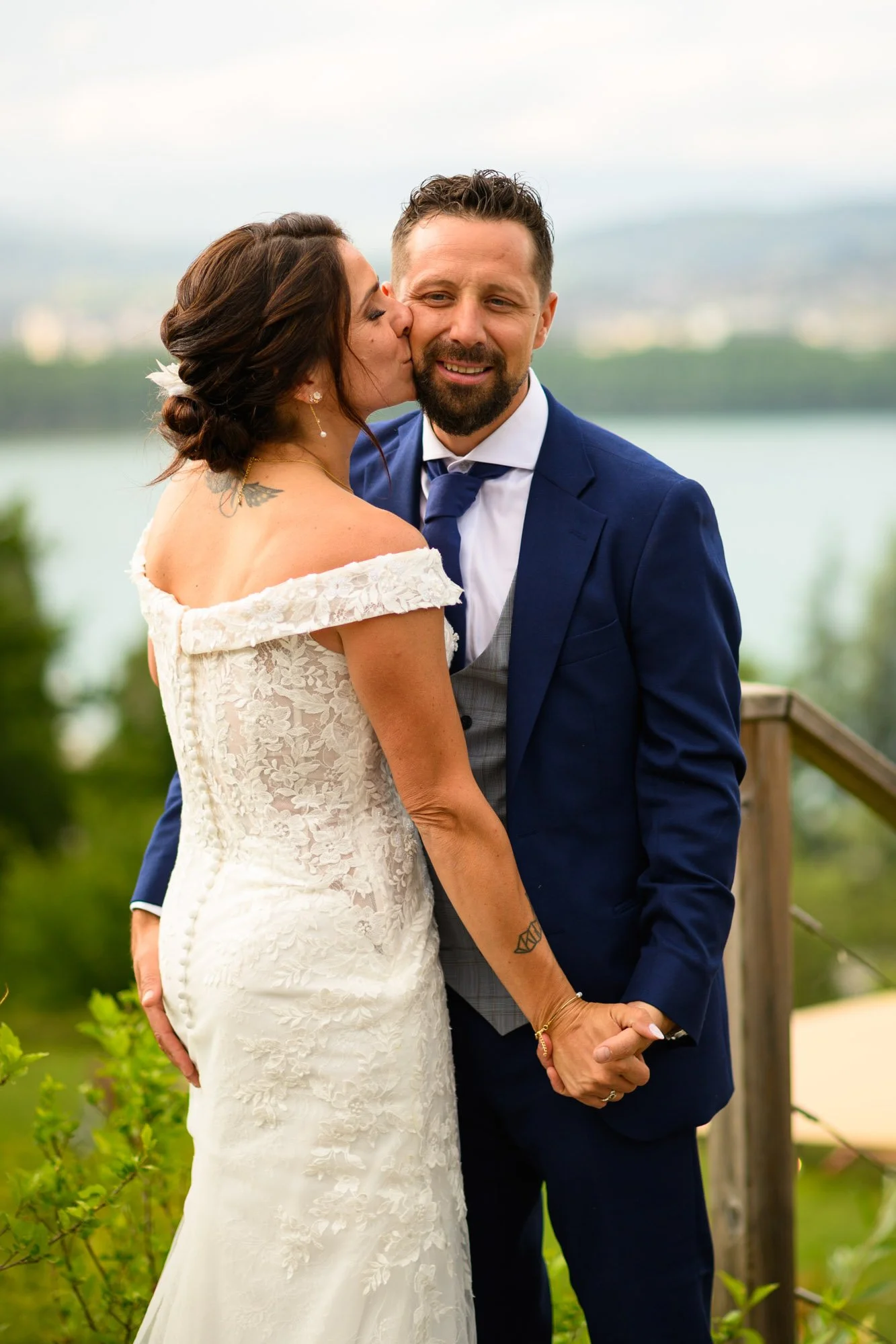 Un couple en mariage se tenant la main, la femme embrassant le mari sur la joue, avec un paysage naturel en arrière-plan.
