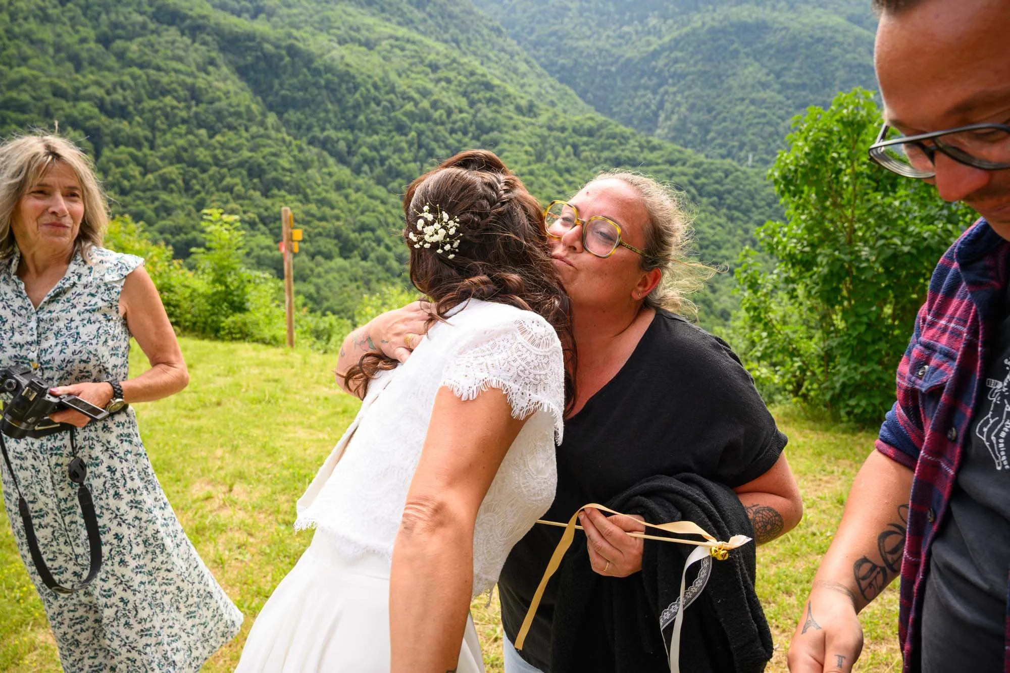 Deux femmes s'embrassent lors d'une cérémonie en extérieur dans un paysage montagneux, une femme porte une robe blanche et des fleurs dans ses cheveux, l'autre porte des lunettes et une tenue noire.