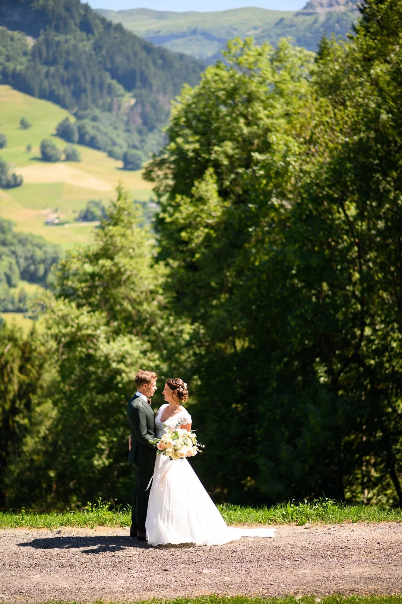 Un couple de mariés se tenant la main devant un paysage verdoyant de montagnes et d'arbres, avec un chemin en premier plan.