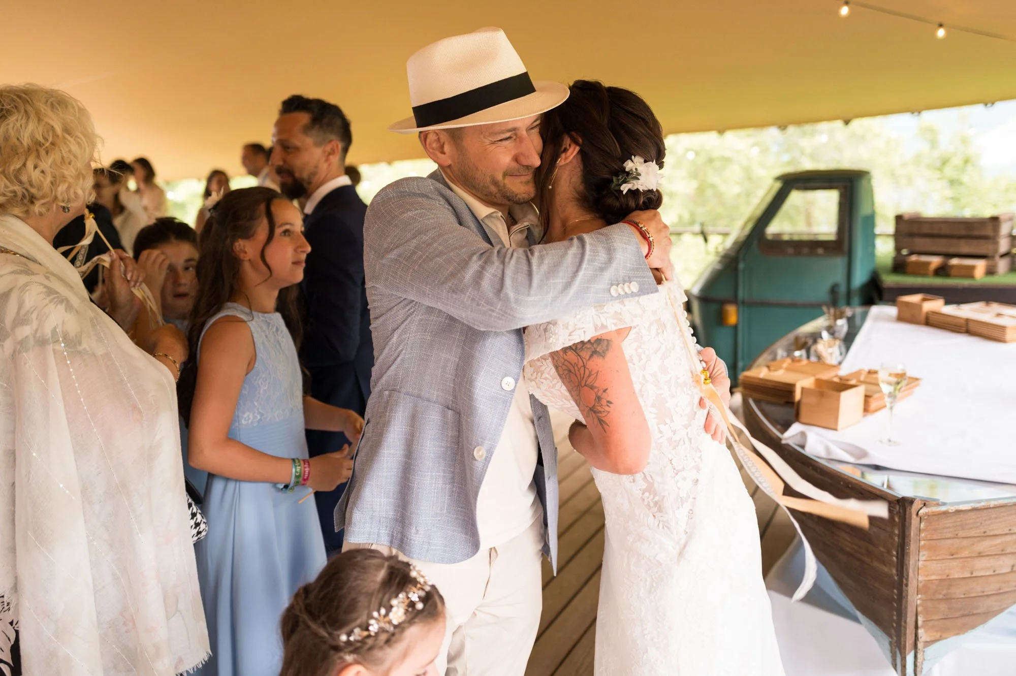 Un homme et une femme s'embrassent lors d'une fête, entourés de plusieurs personnes, sous une tente lors d'un mariage, avec une petite embarcation en bateau à proximité.