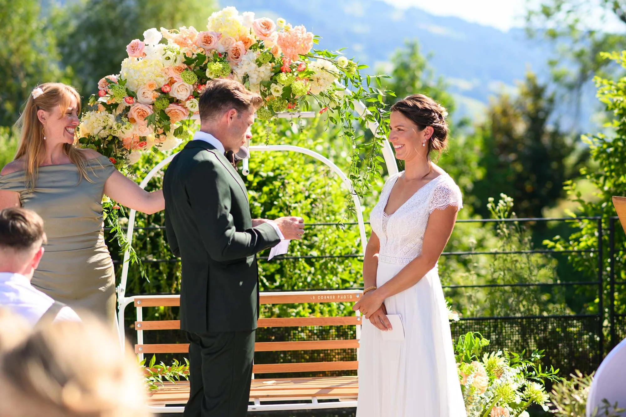 Un mariage en extérieur avec un couple échangeant leurs vœux sous un dais floral, entouré de témoins et d'invités, avec un décor naturel et ensoleillé.