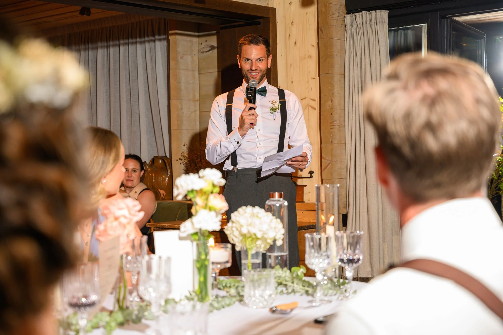 Un homme joue un discours lors d'un mariage, tenant un micro et une feuille, entouré de convives assis à une table décorée de fleurs et de verres.