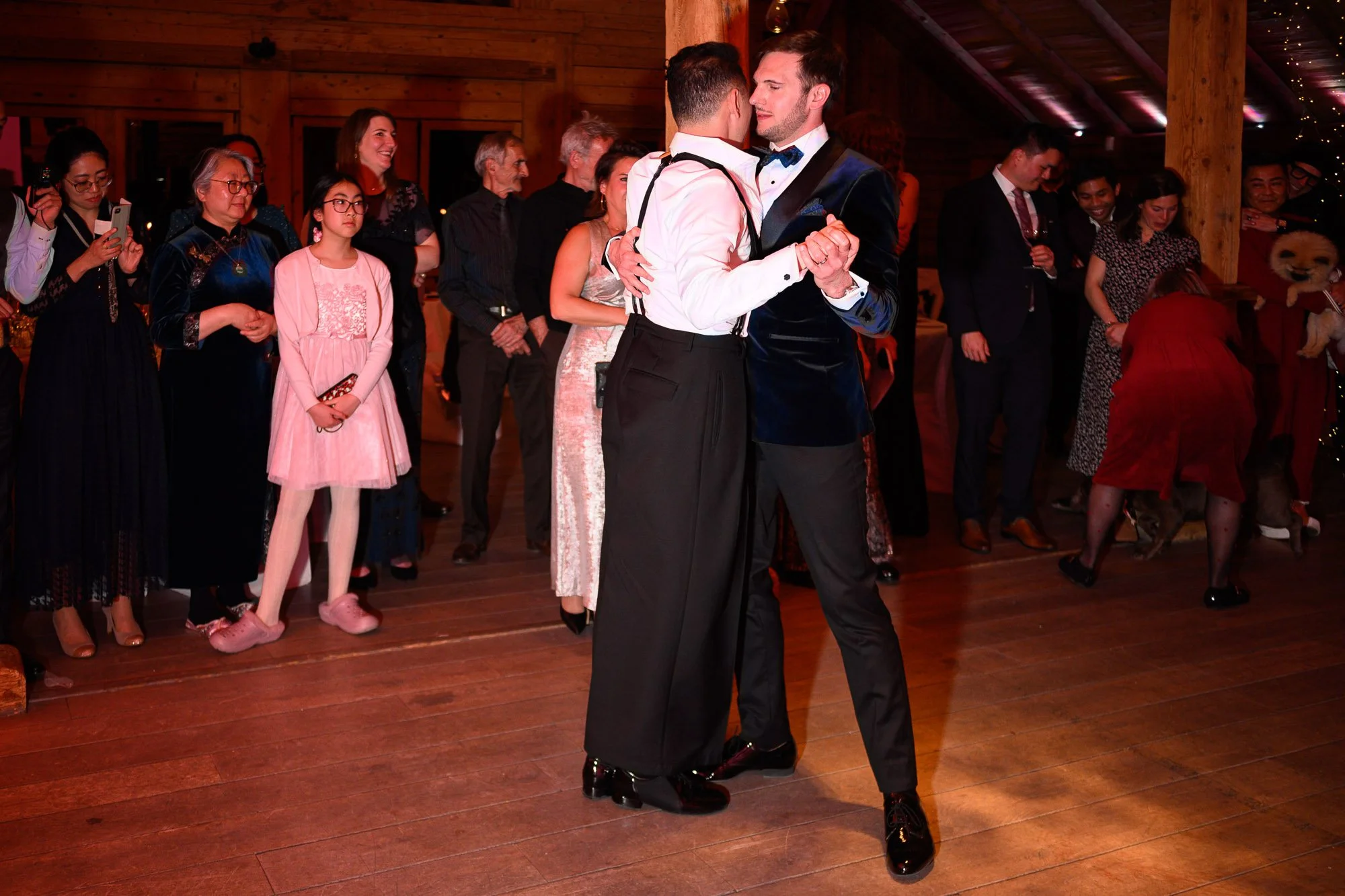 Deux hommes dansant en couple lors d'une fête, entourés de personnes regardant, avec un fond en bois et des lumières. 