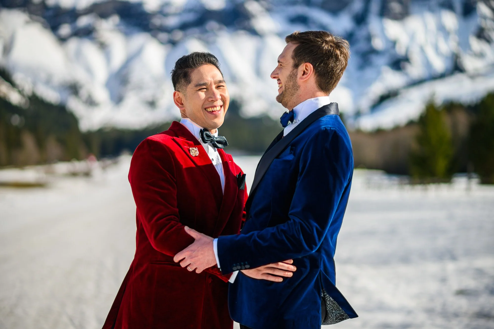 Deux hommes en smokings colorés, un rouge et un bleu, s'embrassent dans un paysage enneigé avec des montagnes en arrière-plan, tous deux souriant.