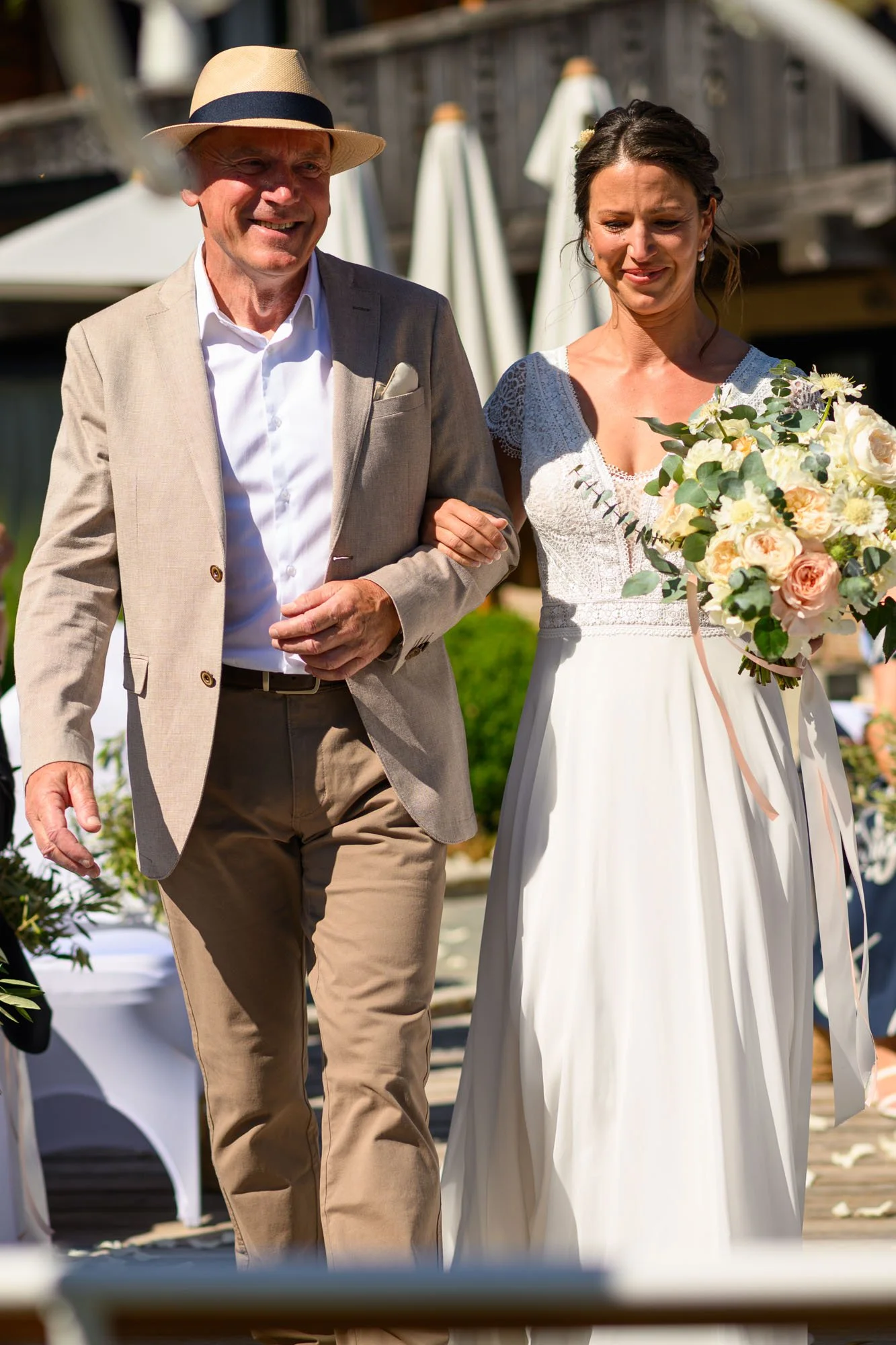 Une jeune femme porte une robe de mariage blanche et tient un bouquet de fleurs, tandis qu'un homme âgé, portant un costume beige, l'accompagne lors d'une cérémonie en plein air.