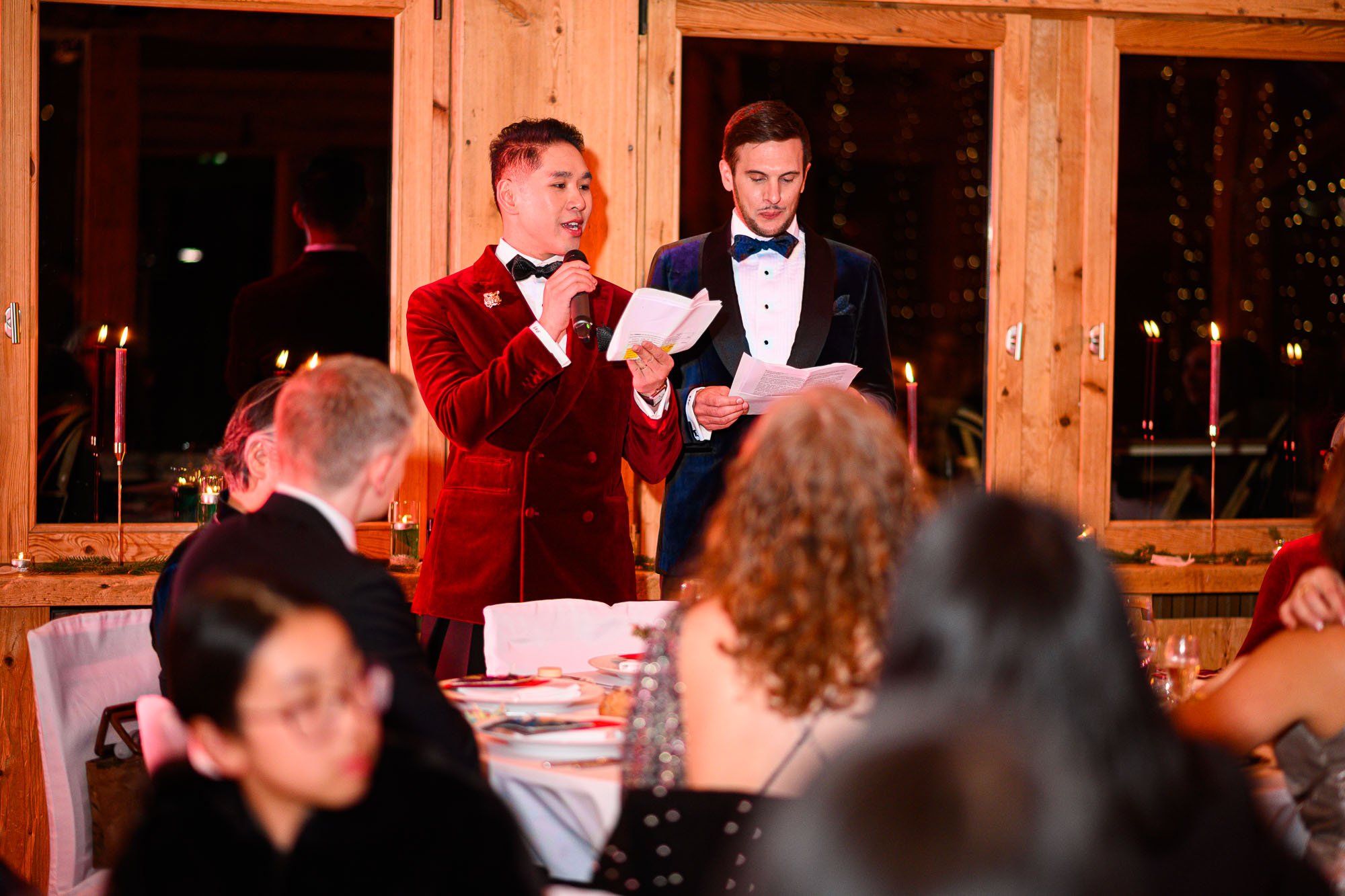 Deux hommes habillés en smoking, l'un en rouge et l'autre en bleu, font un discours lors d'un événement formel dans une pièce en bois avec des bougies, entourés par plusieurs personnes assises à des tables