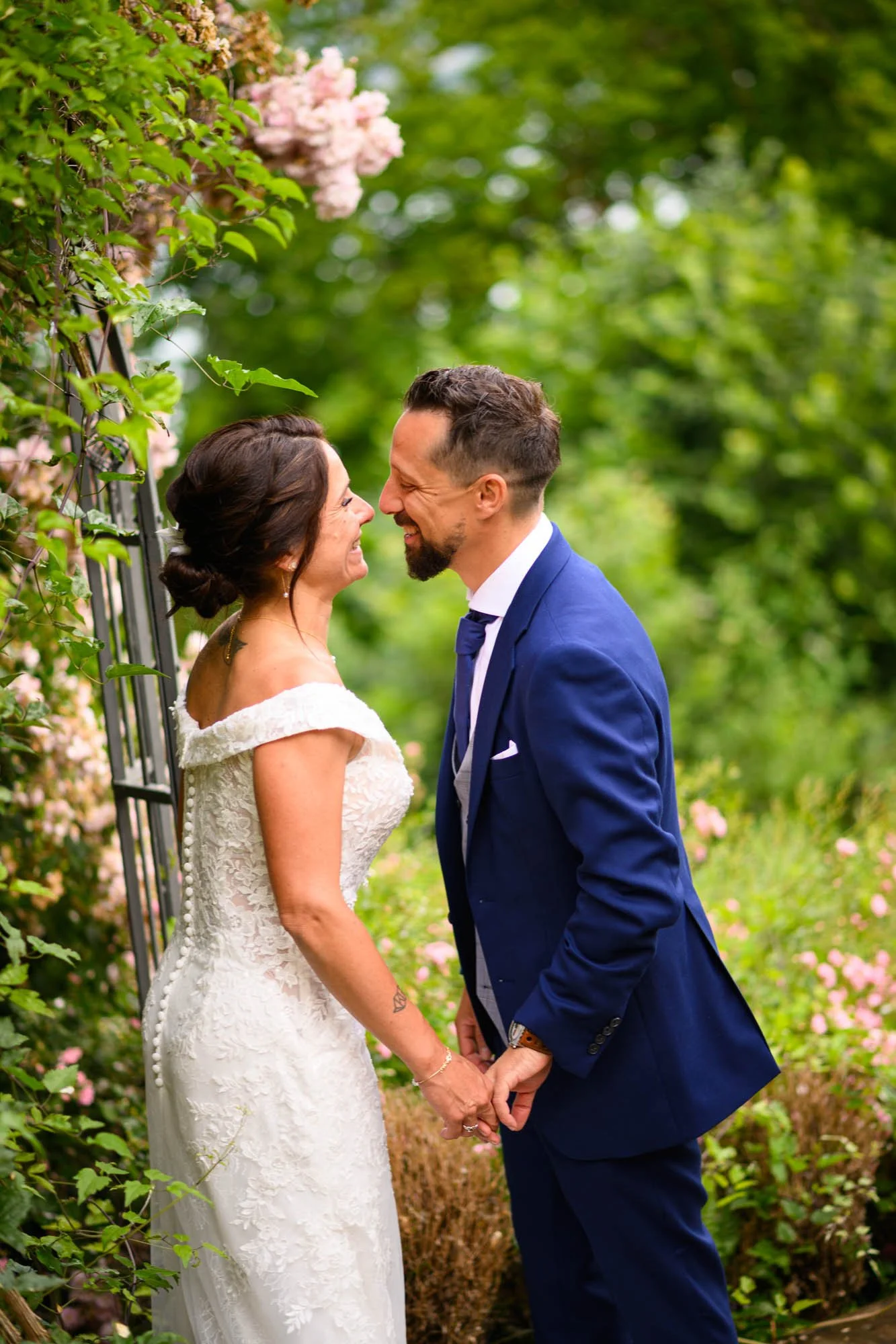 Un couple de mariés se regarde et sourit dans un jardin verdoyant, tenant leurs mains. La mariée porte une robe blanche avec des épaules dénudées, et le marié porte un costume bleu.