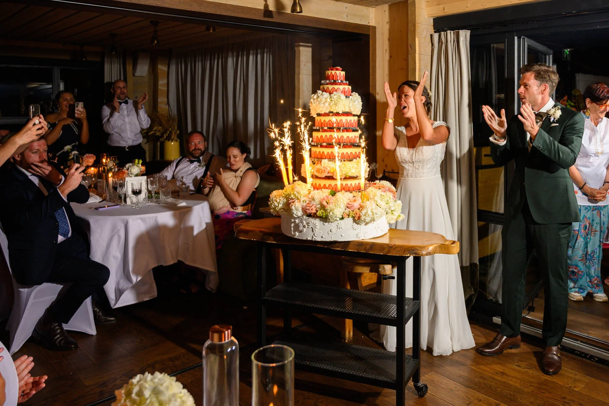 Une femme et un homme, probablement mariés, célébrant le mariage, se tiennent près d'un grand gâteau de mariage décoré de fleurs et de bougies éclairant, entourés d'invités assis et debout dans une salle de réception chaleureuse.
